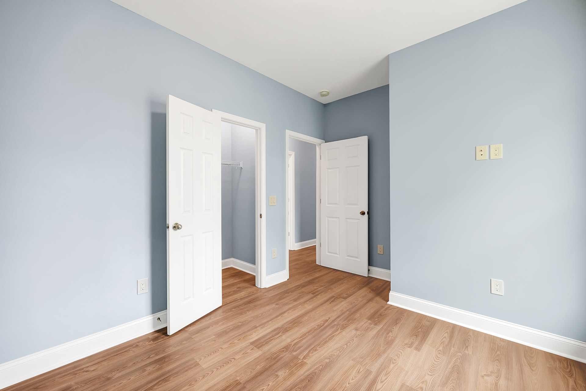 Room with wood flooring, two open white doors featuring gold and silver door knobs, wall outlet with plug, and metal rack against plaster walls.