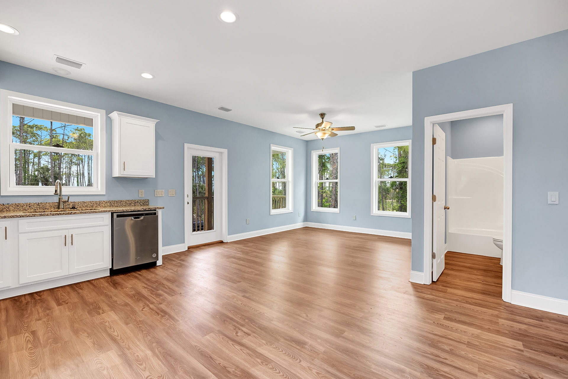 Open kitchen with wood flooring, blue walls, white cabinetry with gold handles, silver dishwasher with black handle, ceiling fan, and window featuring a bird perched on the sill.