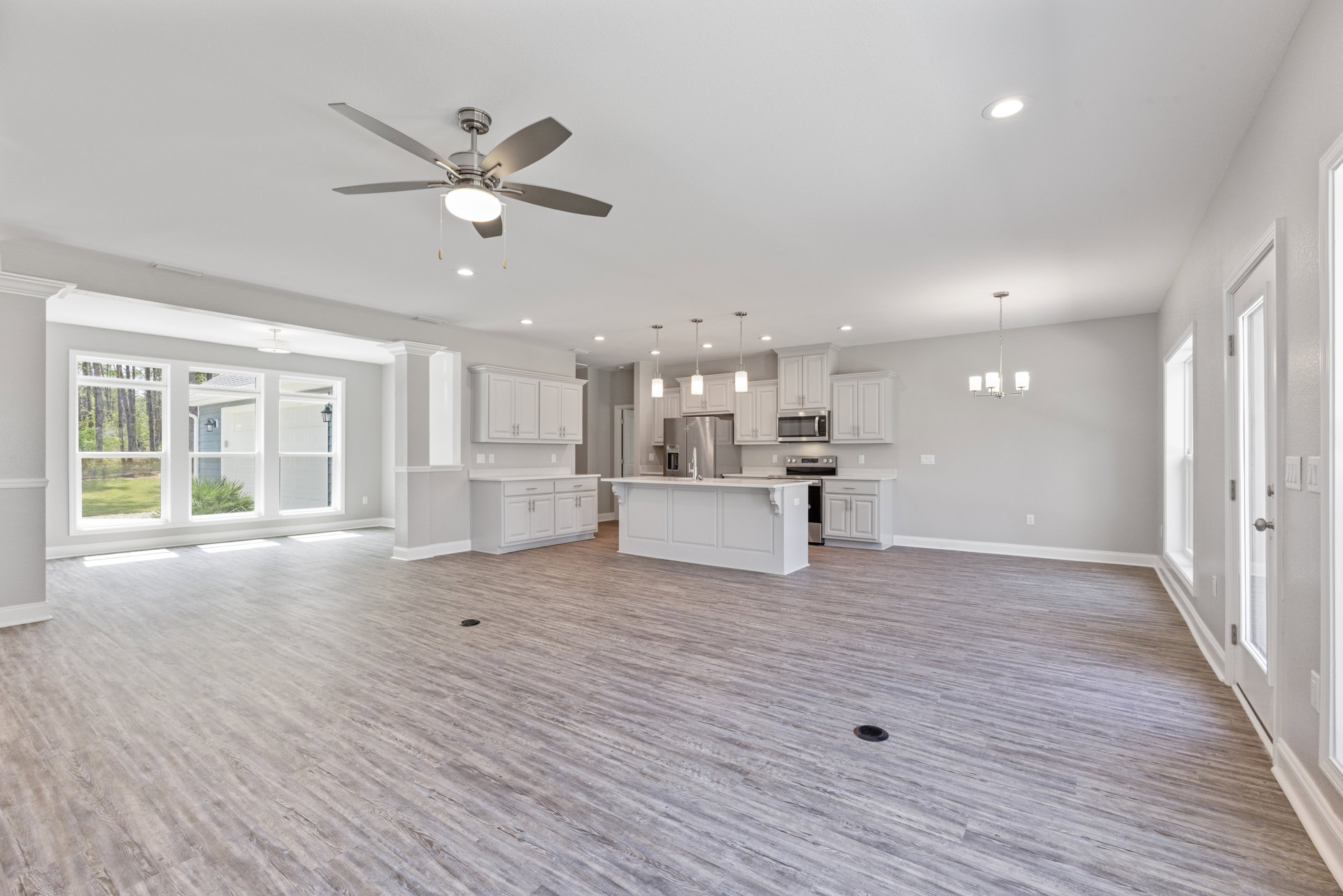 Spacious white living area with wood flooring, ceiling fan with light, open kitchen featuring white countertops and faucet, multiple large windows with views of trees outside