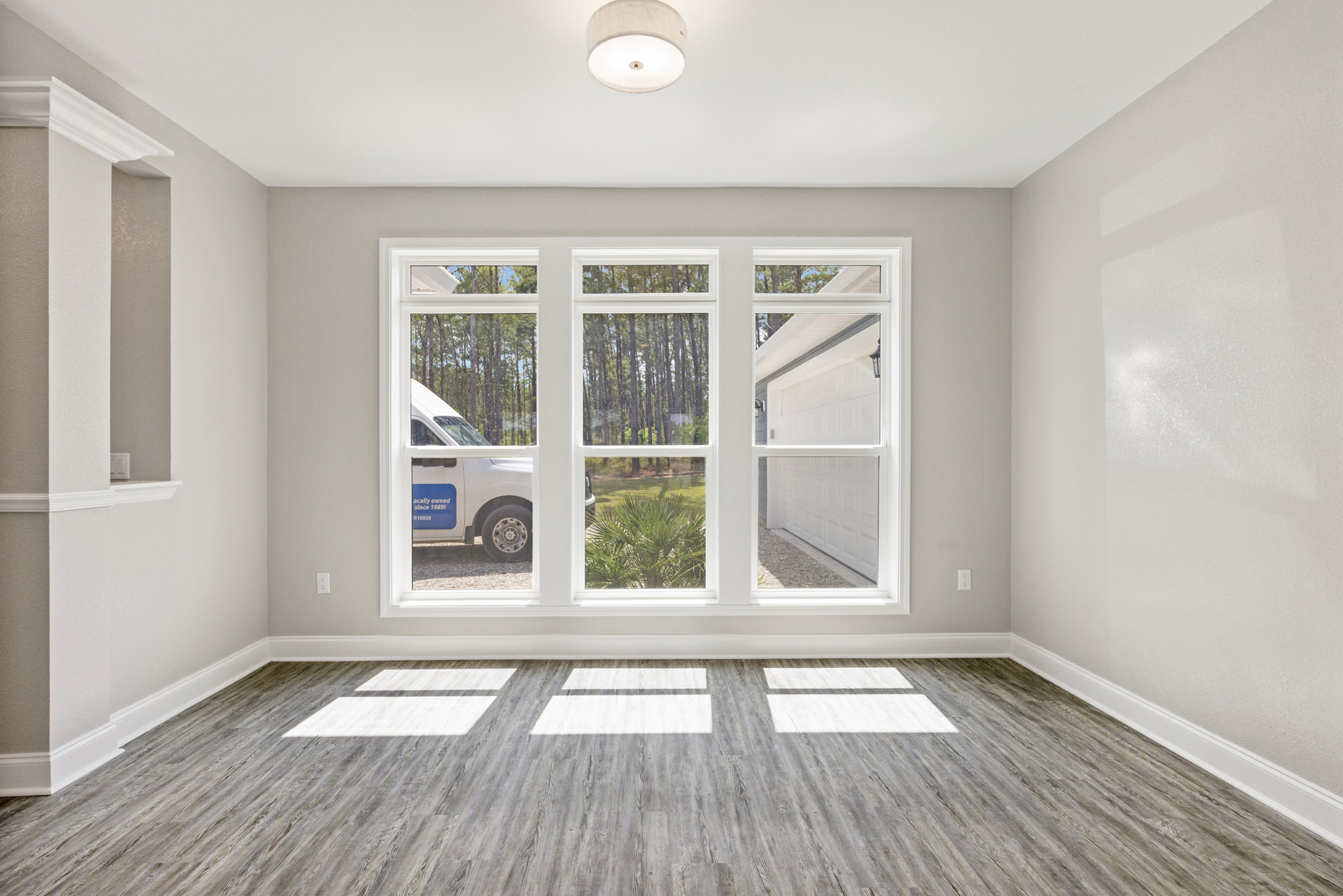 Sunlit room with wide wood plank flooring, large windows framed by white molding, and a modern light fixture; view outside shows a driveway with parked vehicles and leafy trees.