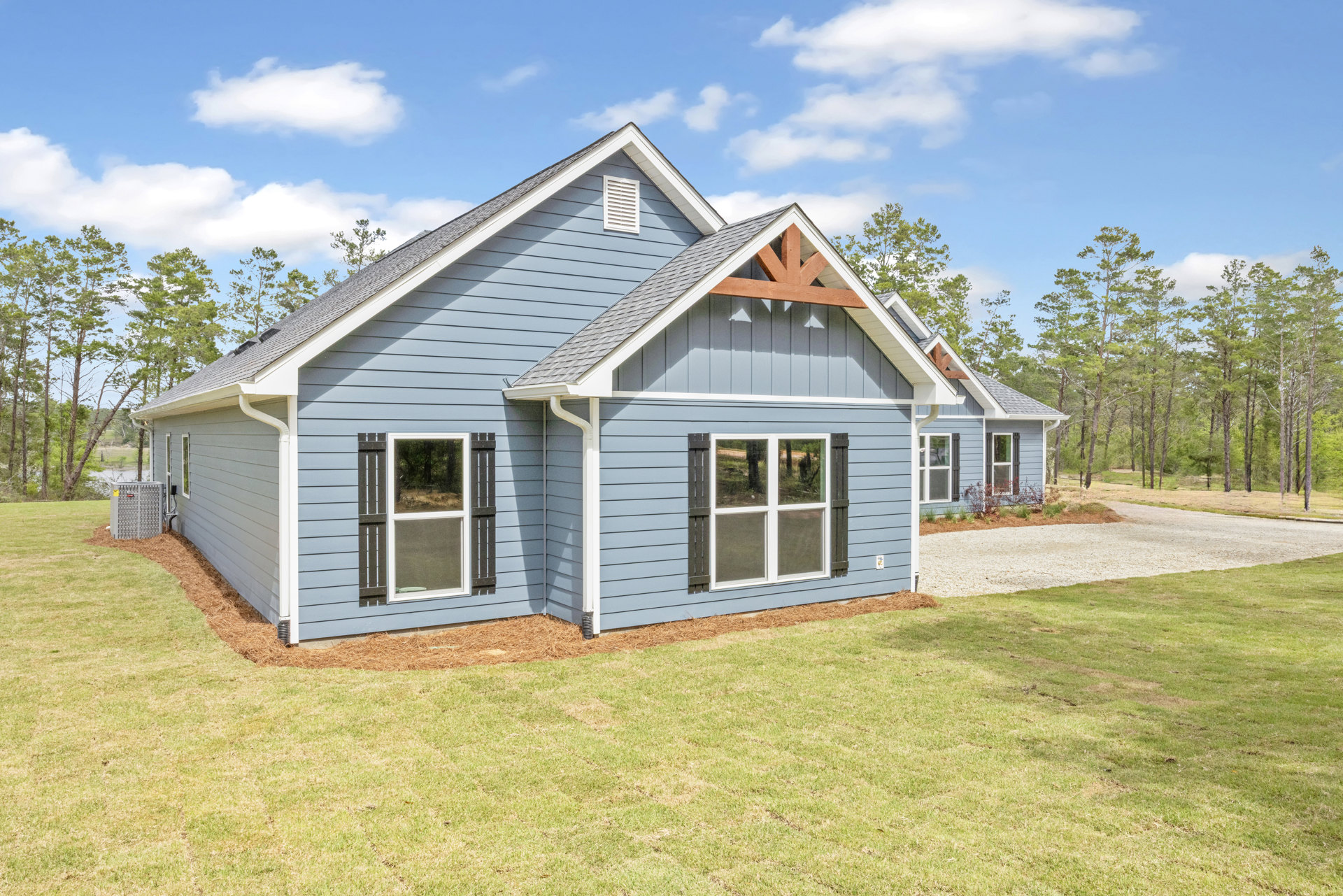 Blue-sided custom home with white trim, blue roof, large front window, concrete driveway, and green lawn bordered by trees under a partly cloudy sky