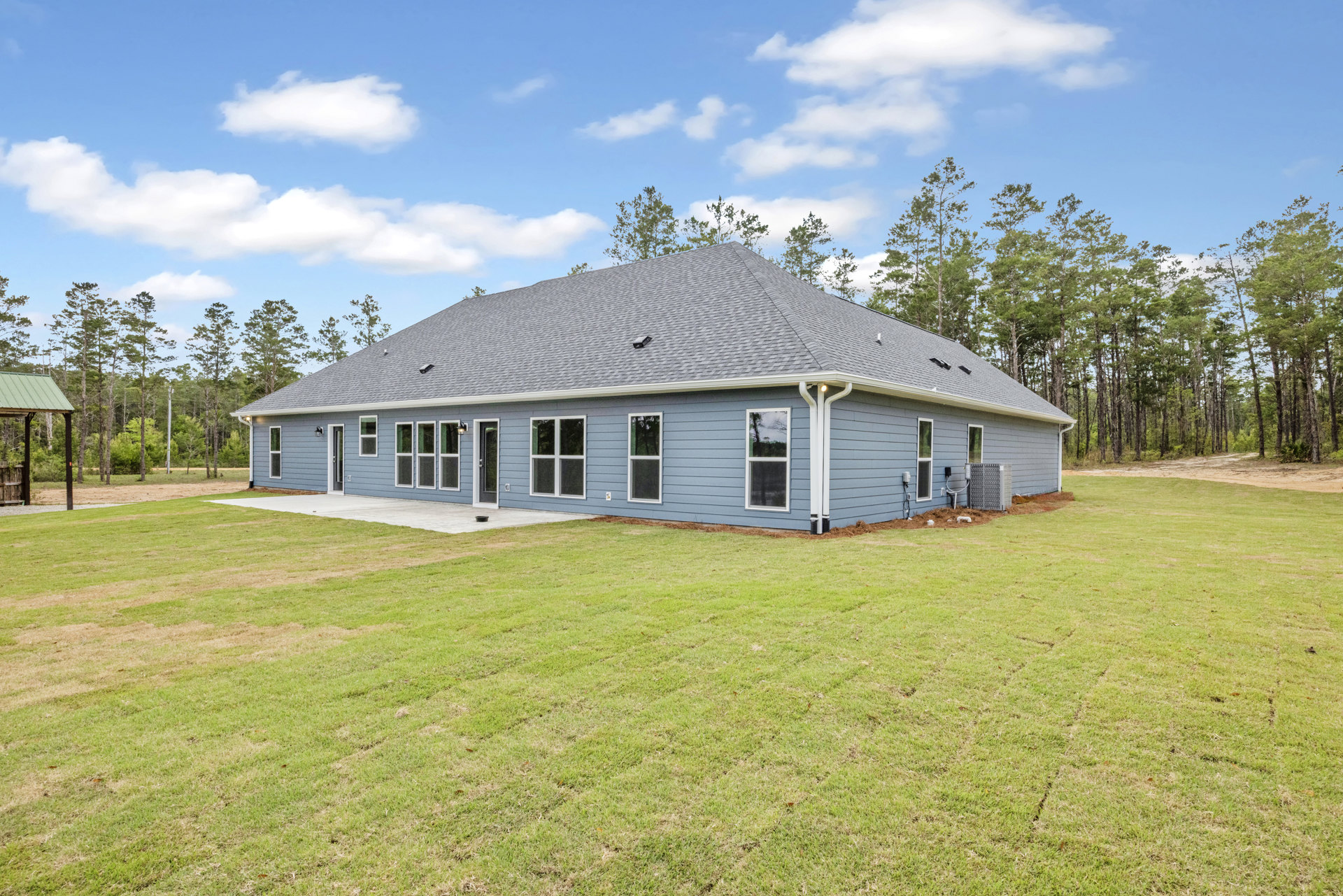 Two-story house with blue roof, white-framed windows, wooden siding, green lawn in front, mature trees in background, partly cloudy sky overhead