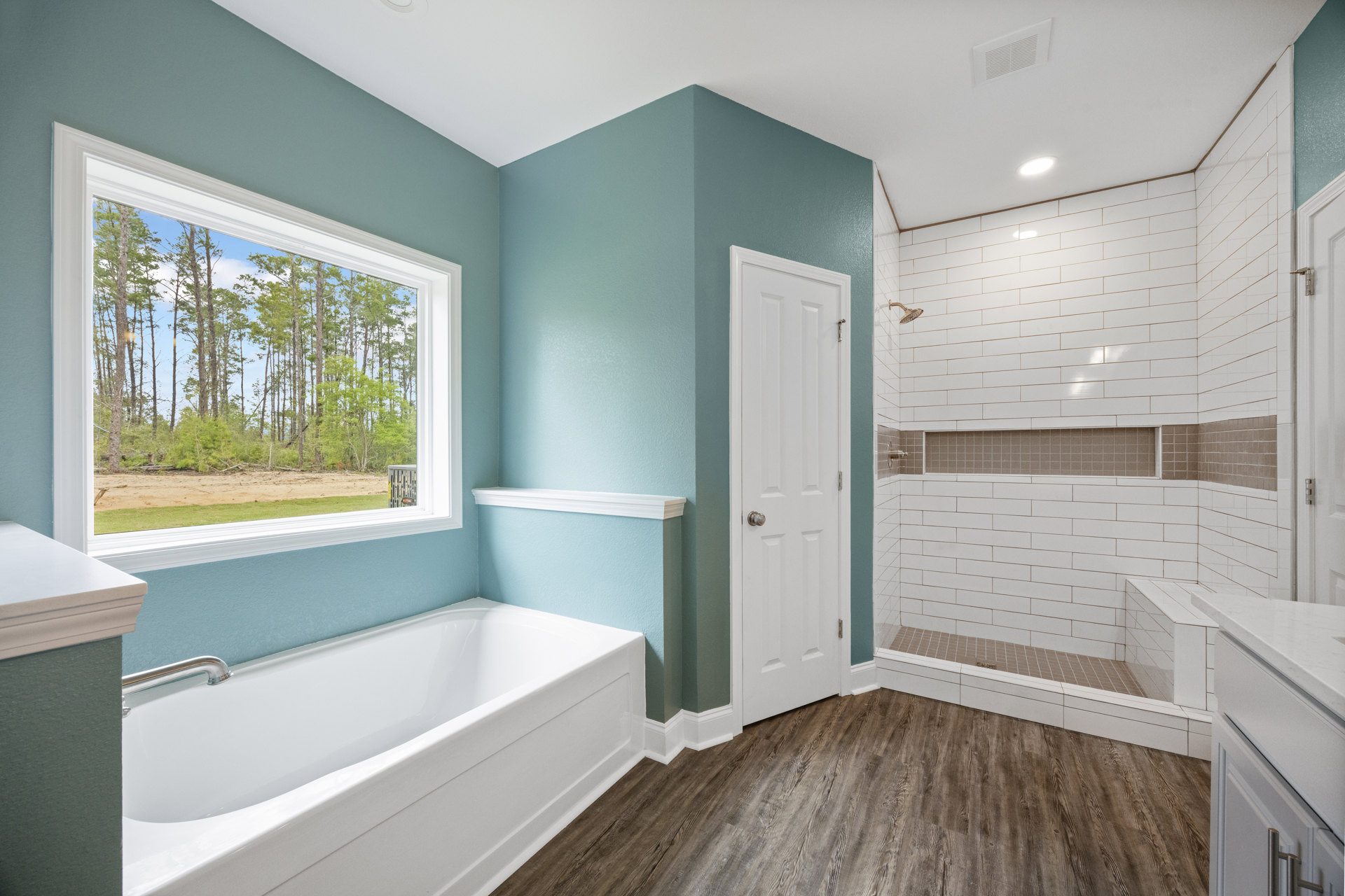 White bathtub with silver faucet beside glass-enclosed shower, light tile flooring, white walls, window showing trees, white door with silver knob