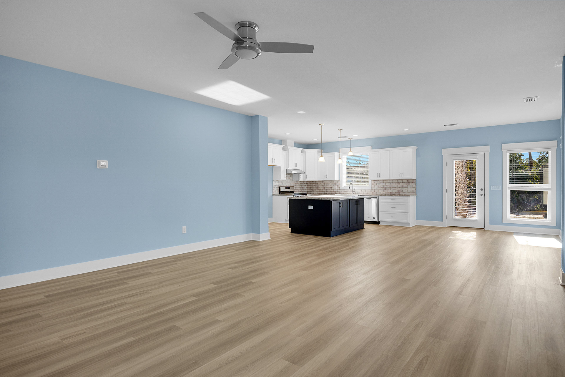 Open-concept room with wood flooring, blue walls, black kitchen island topped with white countertops, ceiling fan with light fixture, palm tree visible through glass door, window