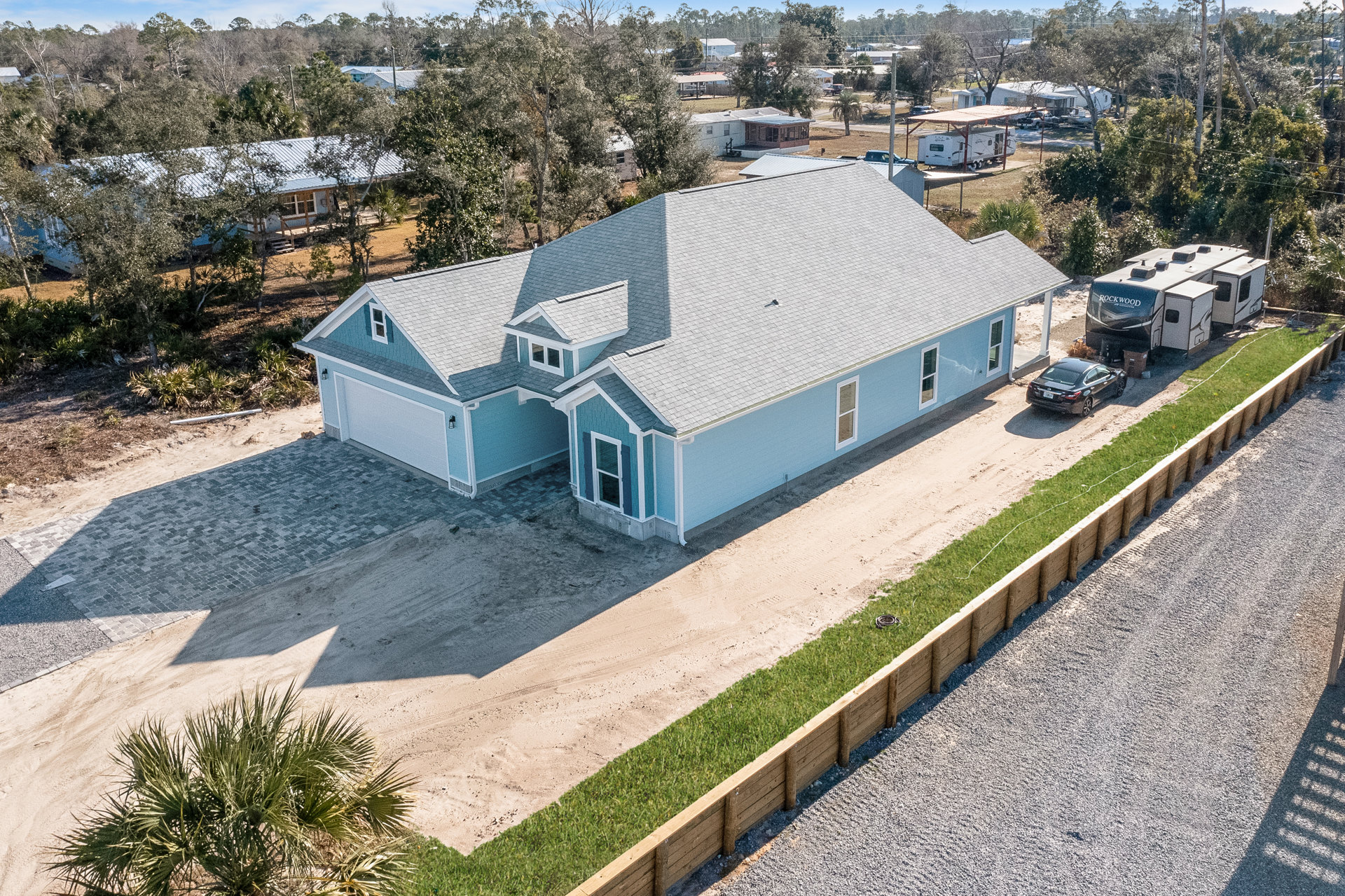 Blue two-story house with white trim, concrete driveway, black car parked in front, palm tree on sandy lawn, large RV beside the home, clear sky overhead