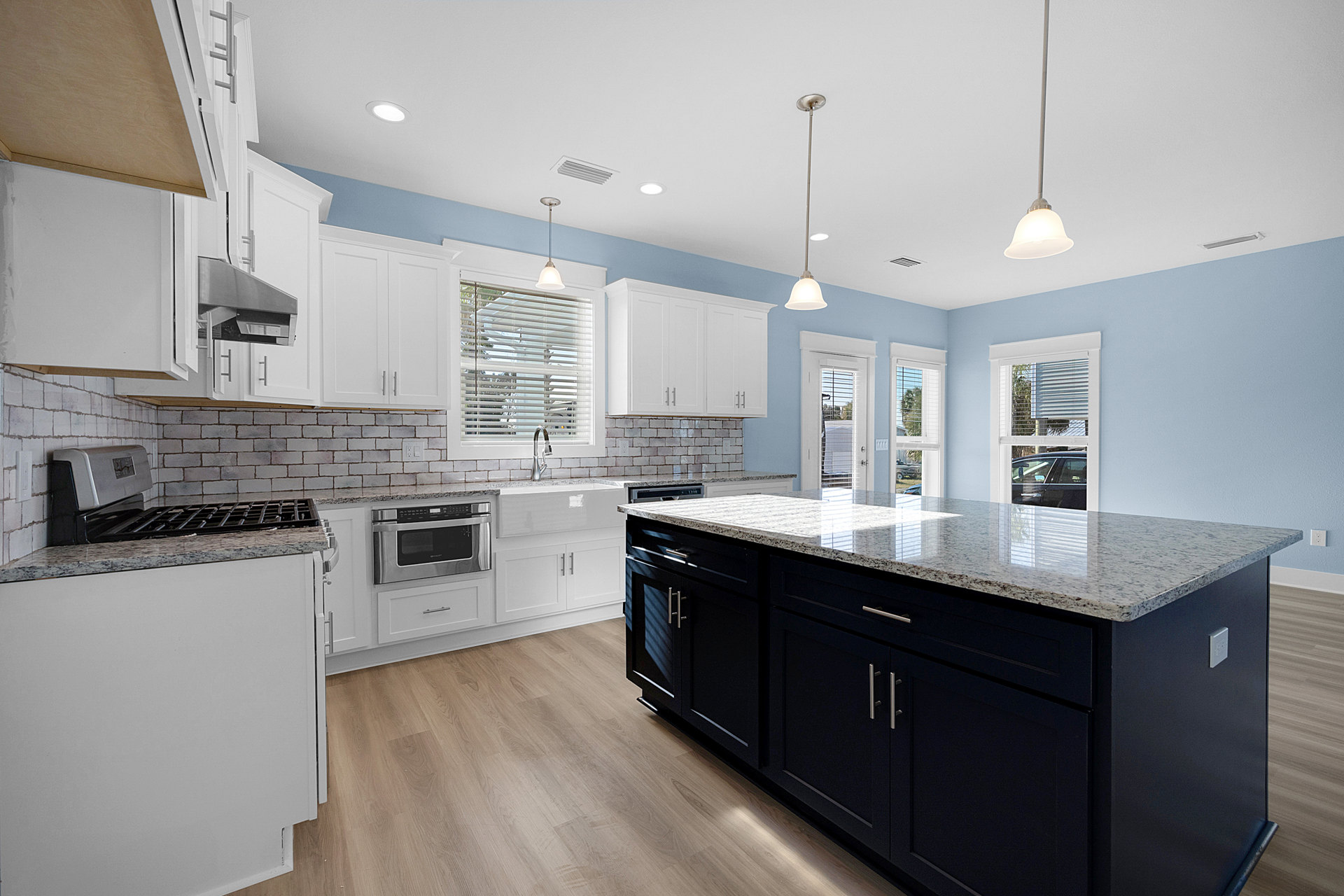 White kitchen cabinets and blue walls, granite island countertop, stainless steel microwave, window with blinds above sink, hardwood flooring