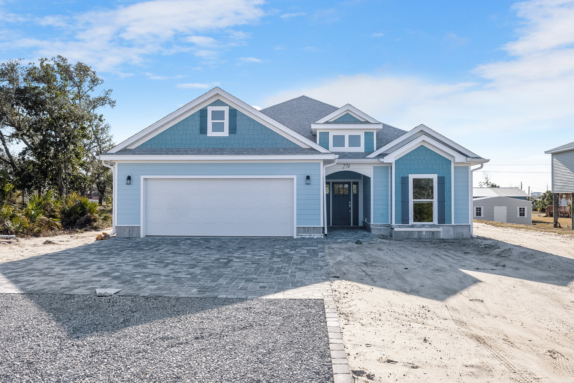 Blue siding exterior with white garage door, paved driveway, white-trimmed windows, and small tree in front yard under partly cloudy sky