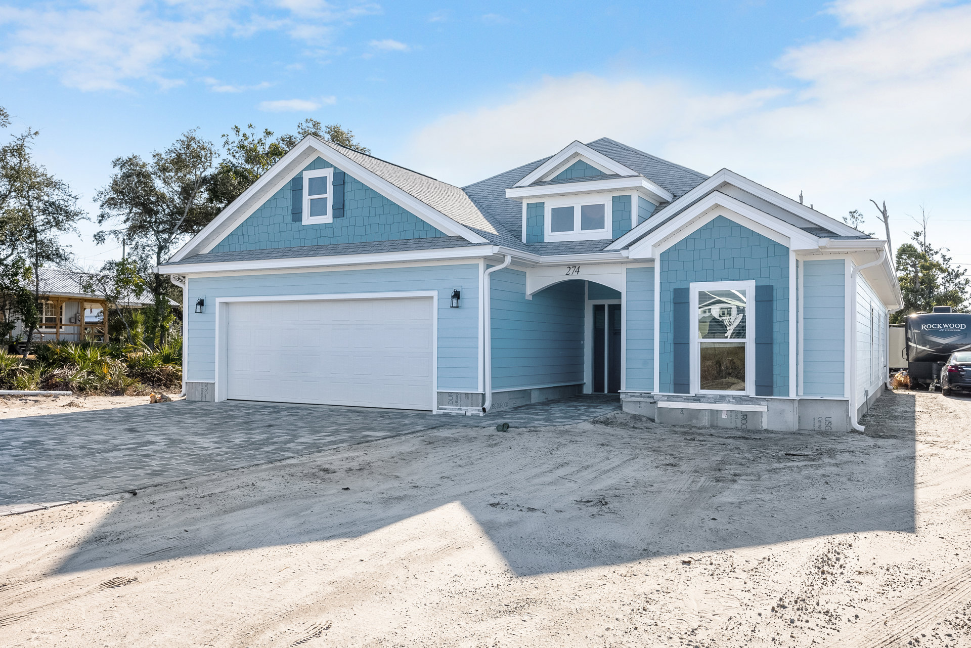 Blue siding house with white garage door, blue roof, front driveway, and ground covered in dirt; broken window visible on facade.