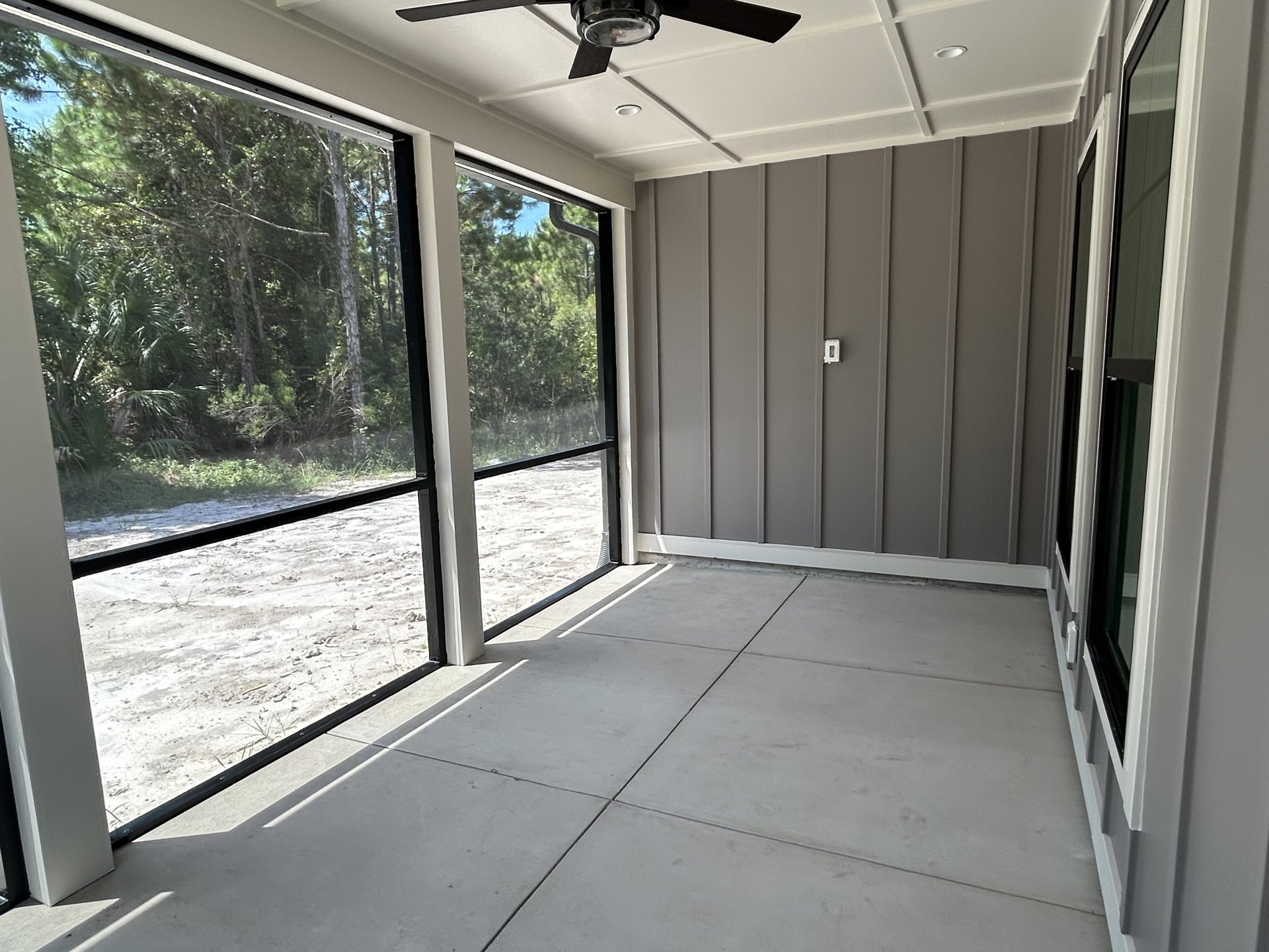 Ceiling fan with light fixture mounted on white ceiling, large window framed in black overlooking green trees, white tile flooring, natural daylight filling the room