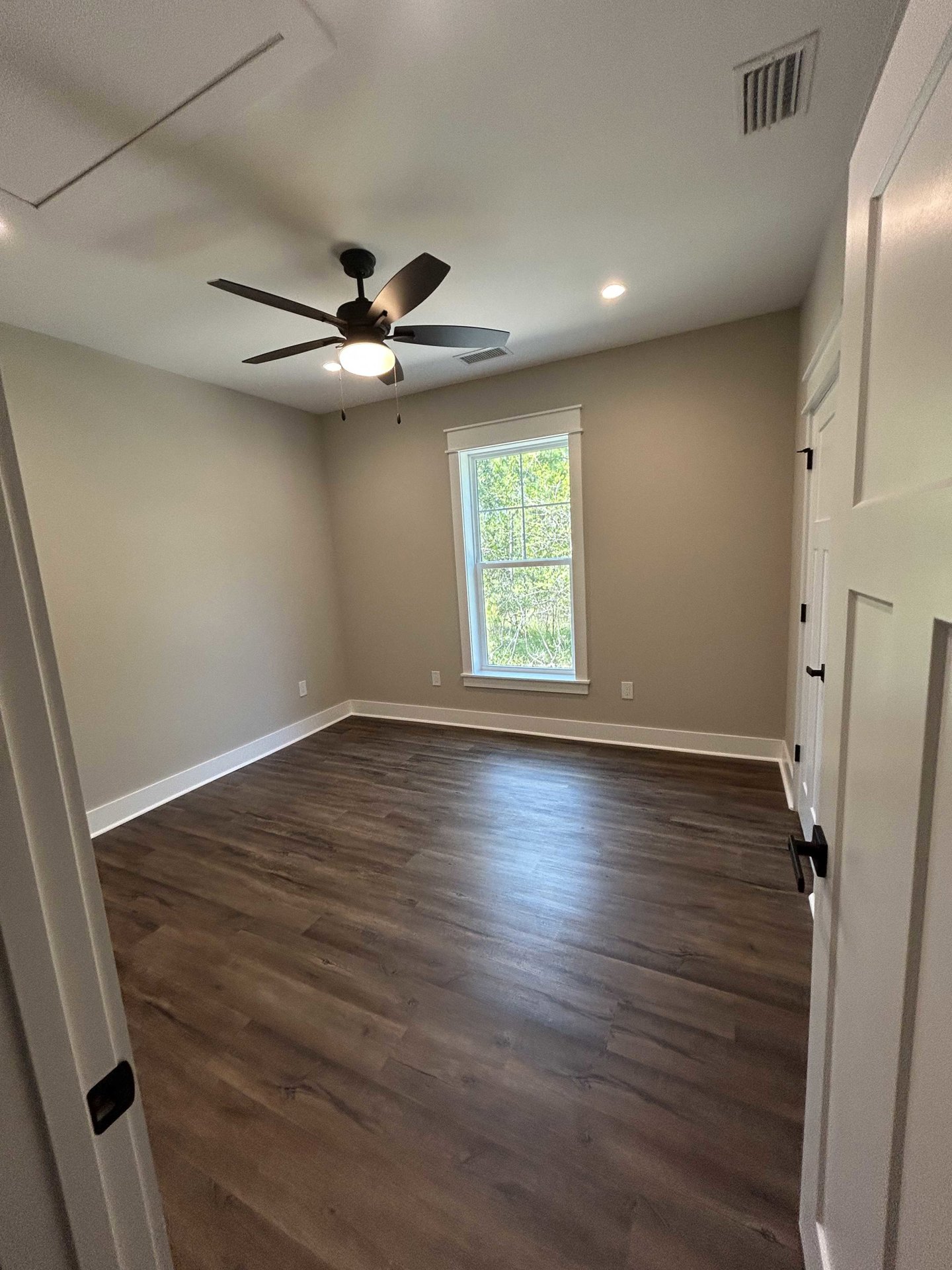 Hardwood floor room with white walls, ceiling fan with light, large window showing green trees, wall vent near baseboard