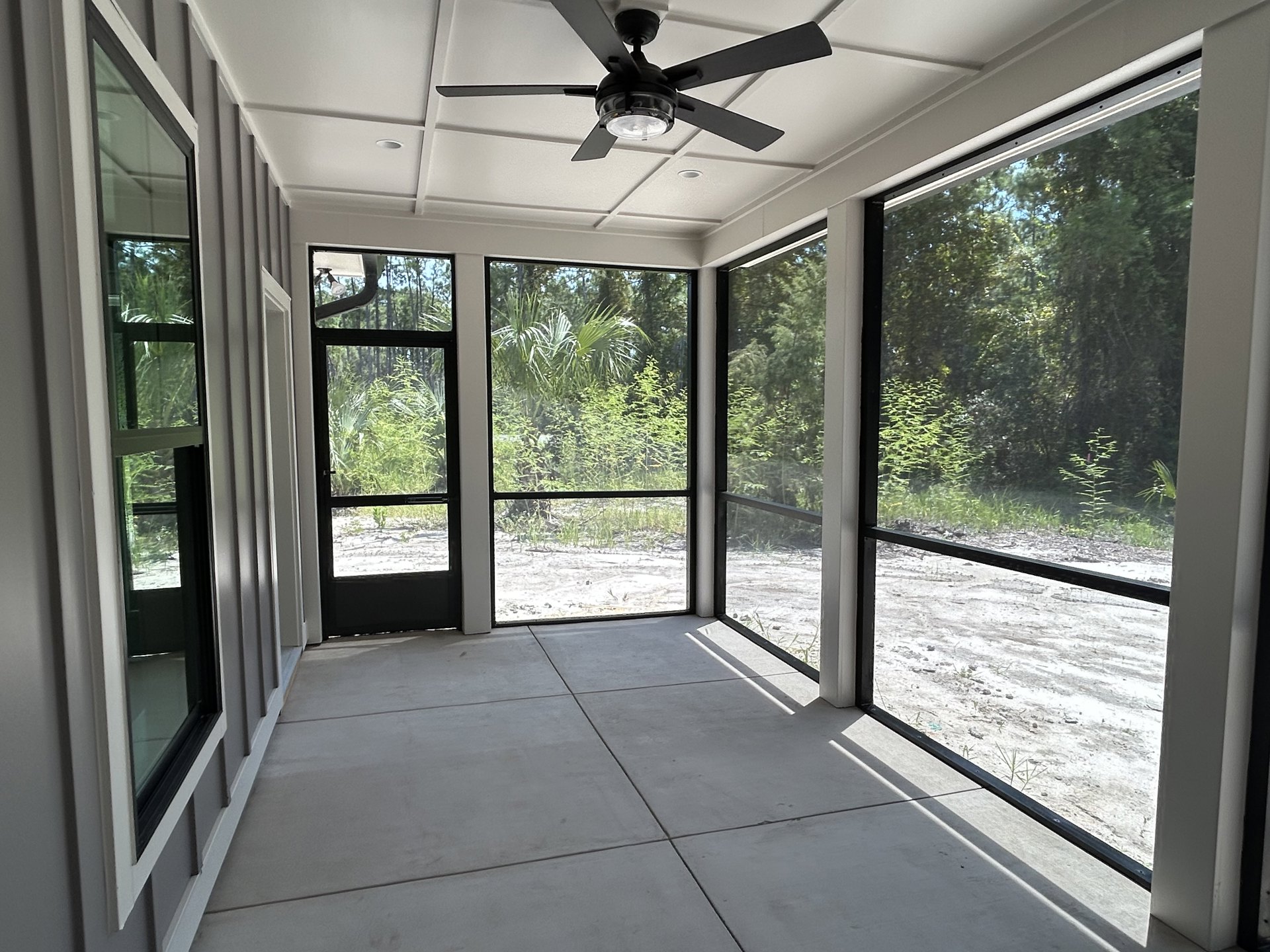 Ceiling fan with light fixture above white tile floor, large glass windows revealing leafy outdoor plants, neutral walls, and daylight streaming into spacious room.