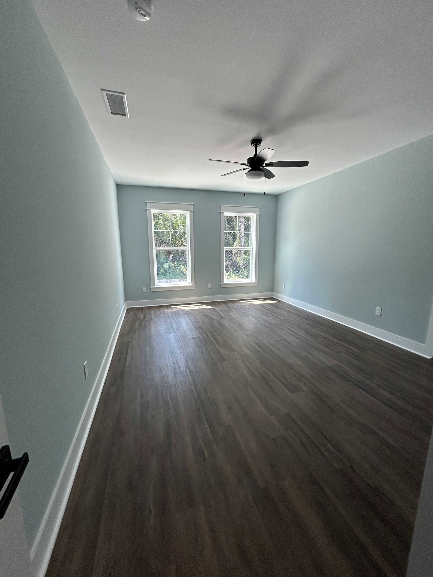 Room with dark wood flooring, white plaster walls, ceiling fan with light fixture, large window overlooking trees.