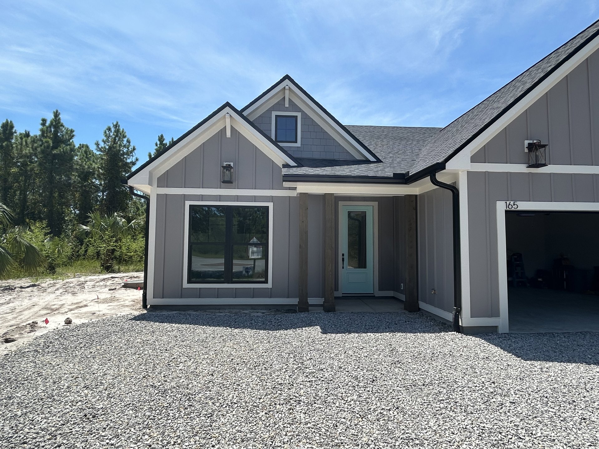 Two-story home with light siding, black-framed windows, gravel driveway, and mature trees and bushes in the background
