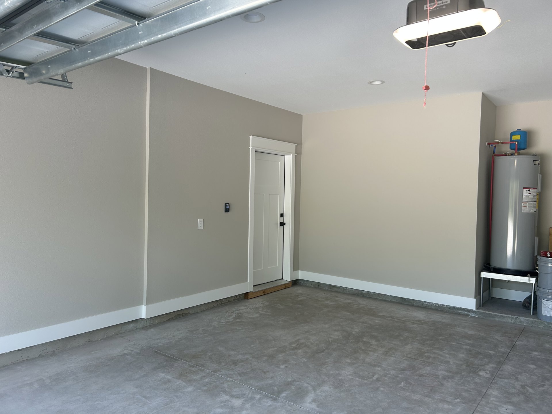Concrete-floored utility room with white door featuring black handle, white water tank connected to red pipe, blue container with yellow label, and ceiling-mounted light fixture