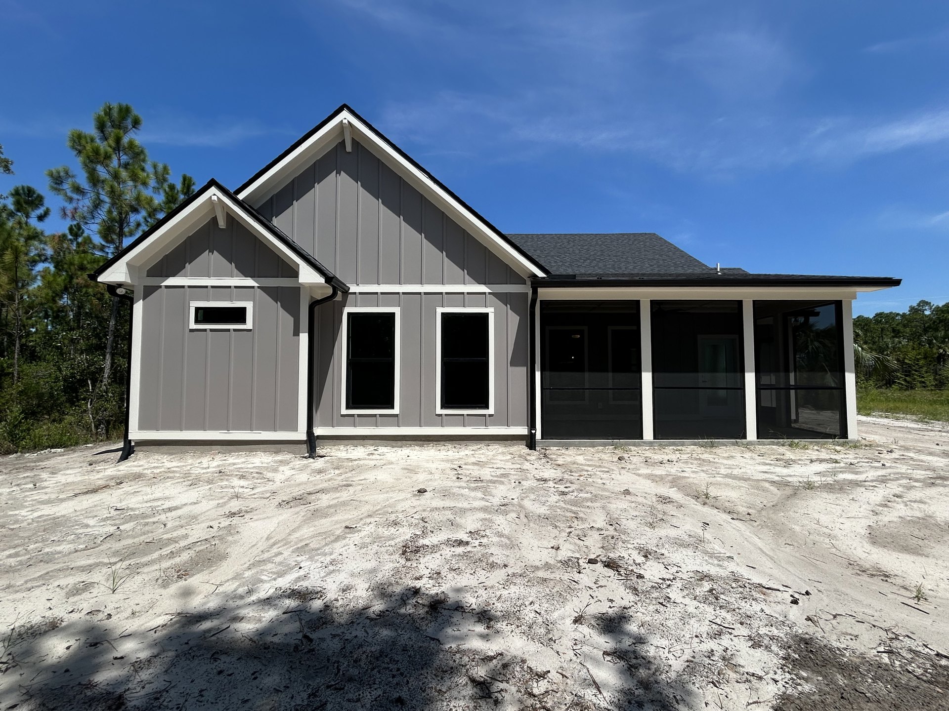 Two-story house with black roof, black-framed windows, and black front door, white siding, gravel driveway leading to attached garage, mature trees in background, cloudy sky