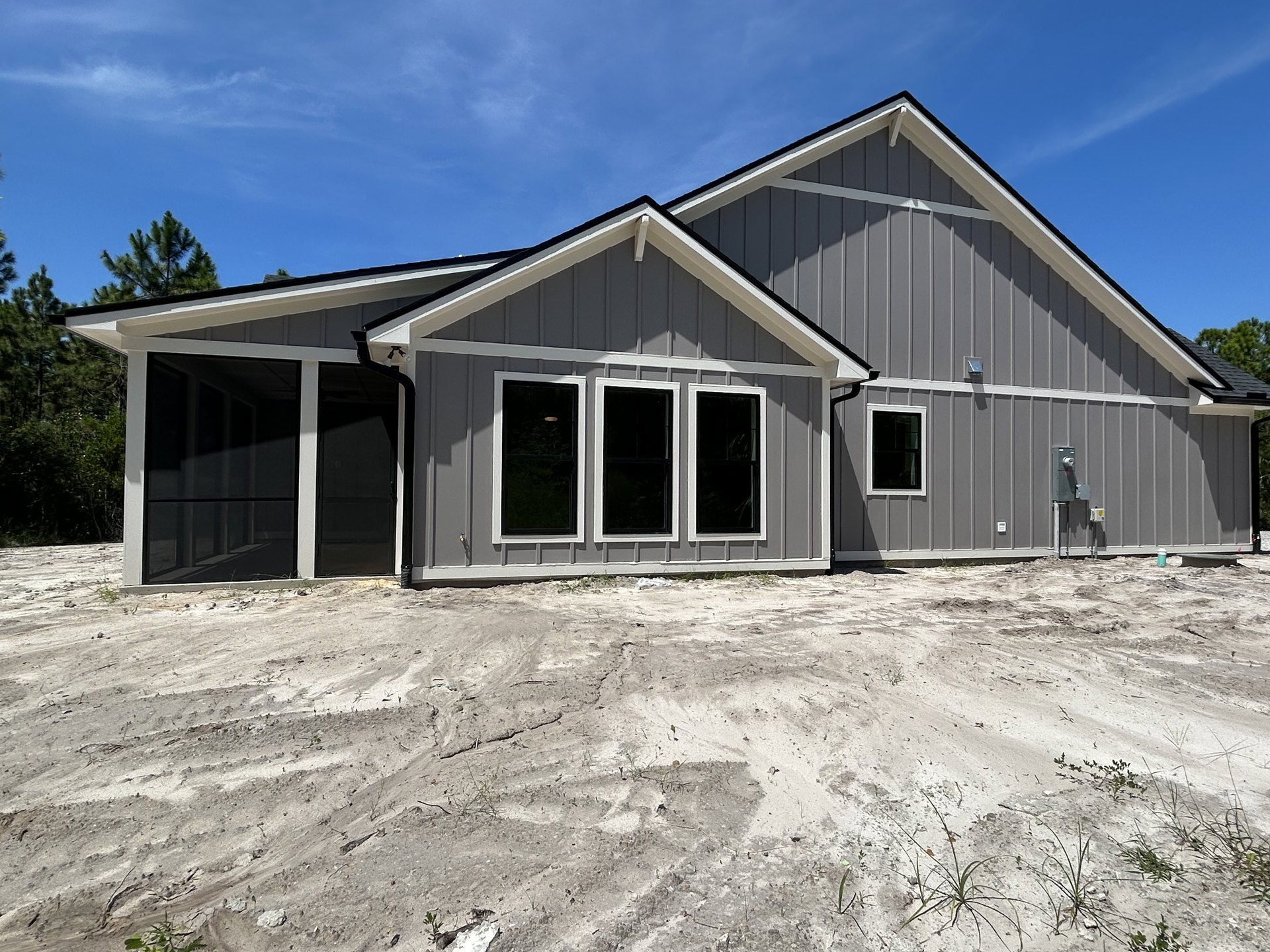 Modern home exterior with black-framed windows, light visible inside, surrounded by bare dirt and minimal landscaping under a cloudy sky