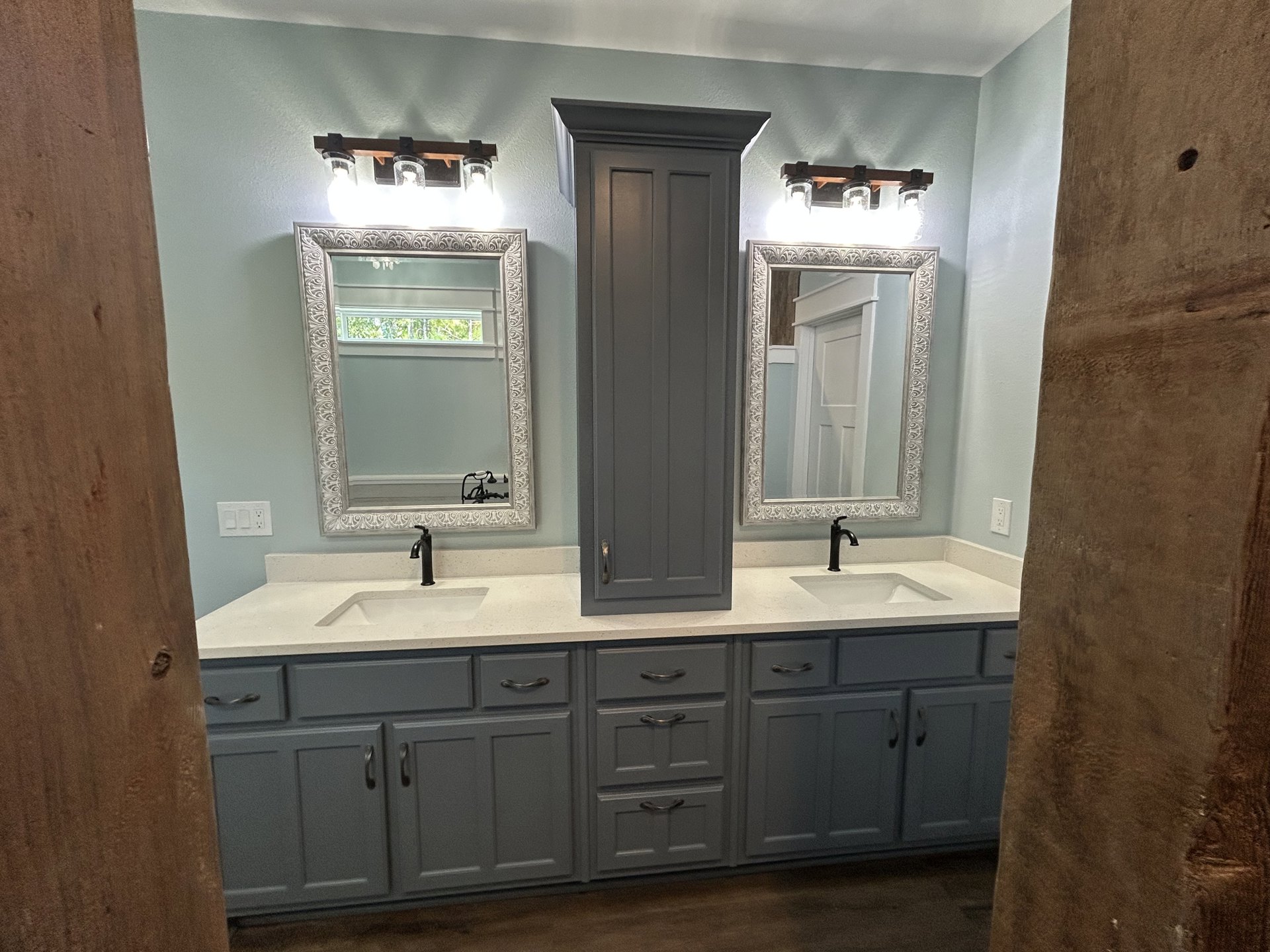Bathroom with two undermount sinks set in a stone-topped vanity, framed mirrors above each sink, white paneled door, wood cabinetry, chrome faucets, and a window providing natural