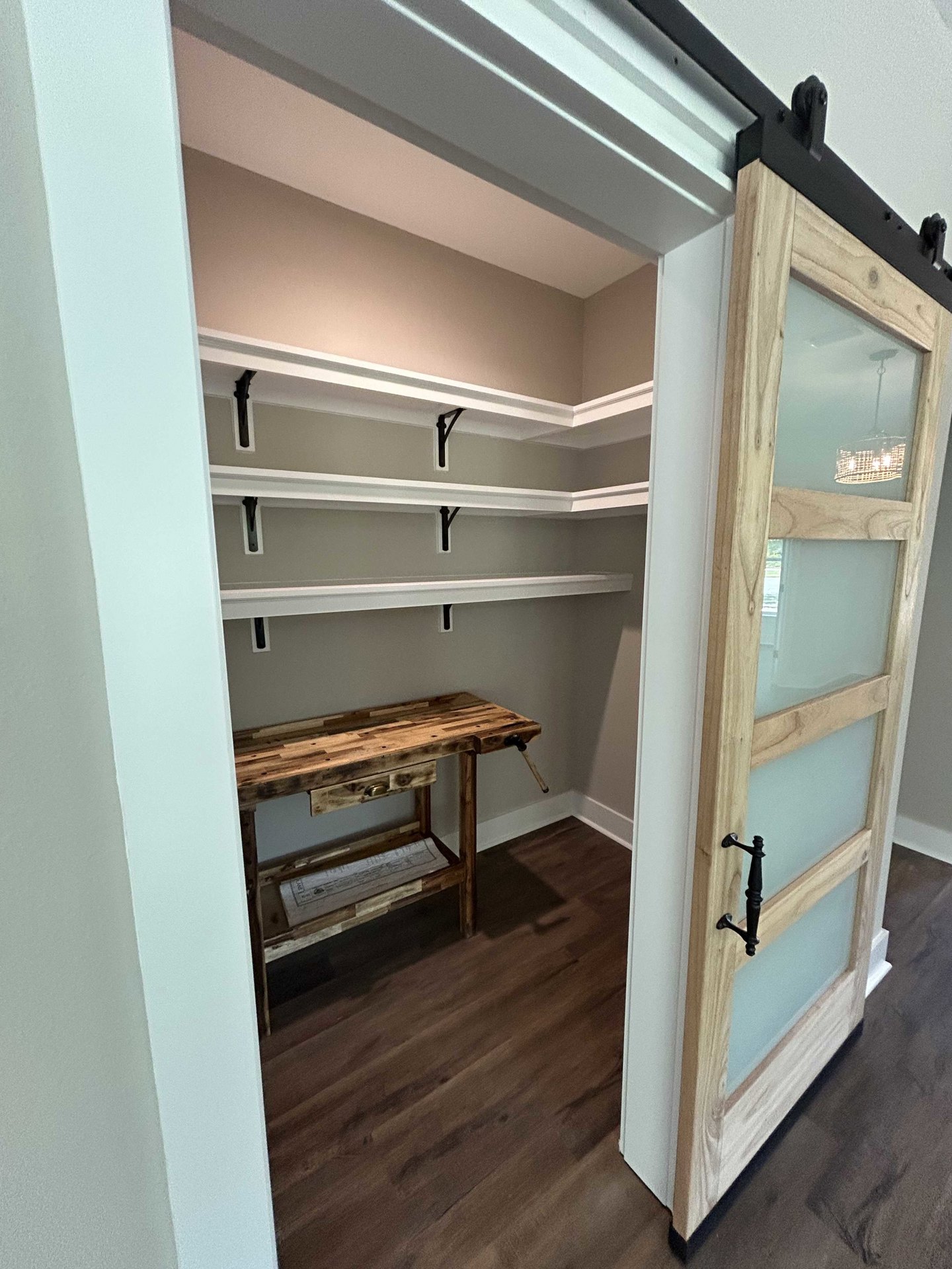 Walk-in closet with built-in plywood shelves, hardwood floor, wooden desk with drawer, frosted glass door, and black decor item on desk