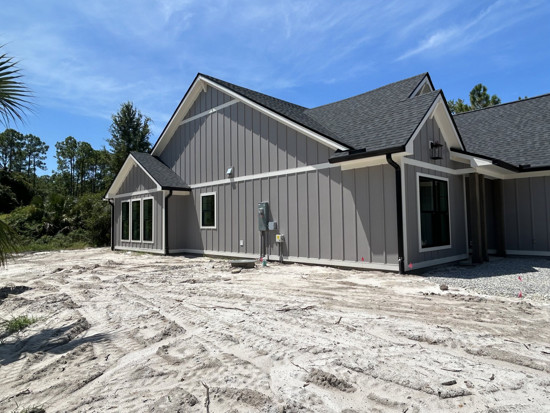 Modern home under construction featuring black-framed windows, grey exterior walls, metal fencing, exposed pipes, and a partially finished roof, surrounded by mature trees.