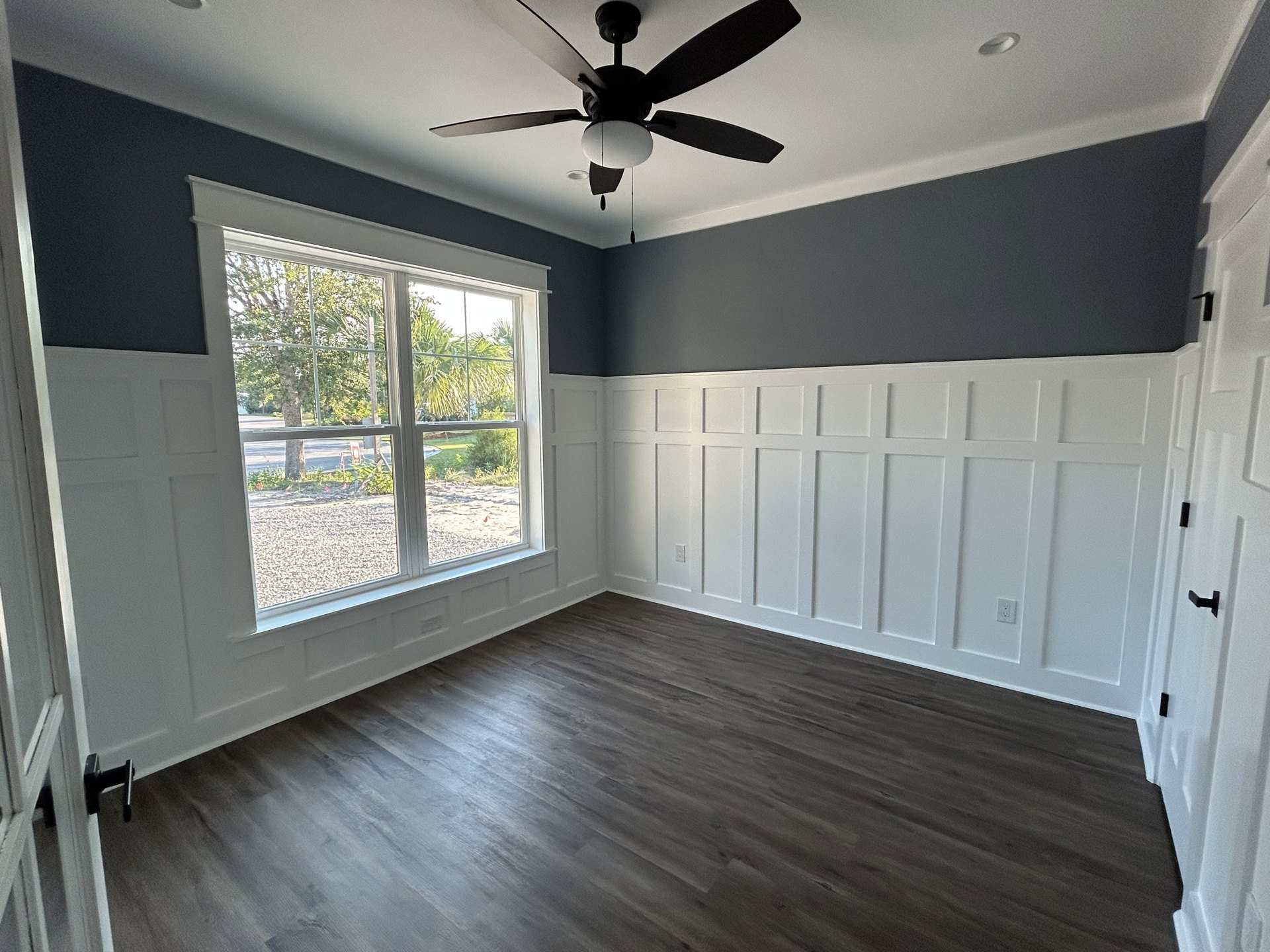 Ceiling fan with light fixture above wood floor, white paneled walls, window overlooking street and trees