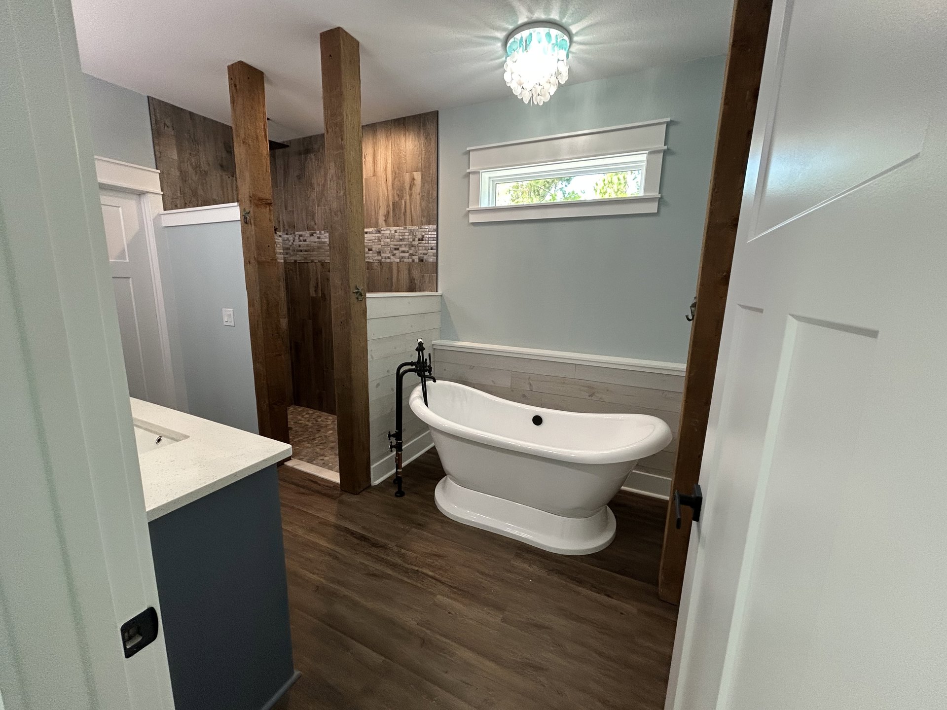 Freestanding white tub with black drain button on wood plank floor, white-framed window, modern light fixture, chrome door handle, and white toilet against light-colored walls