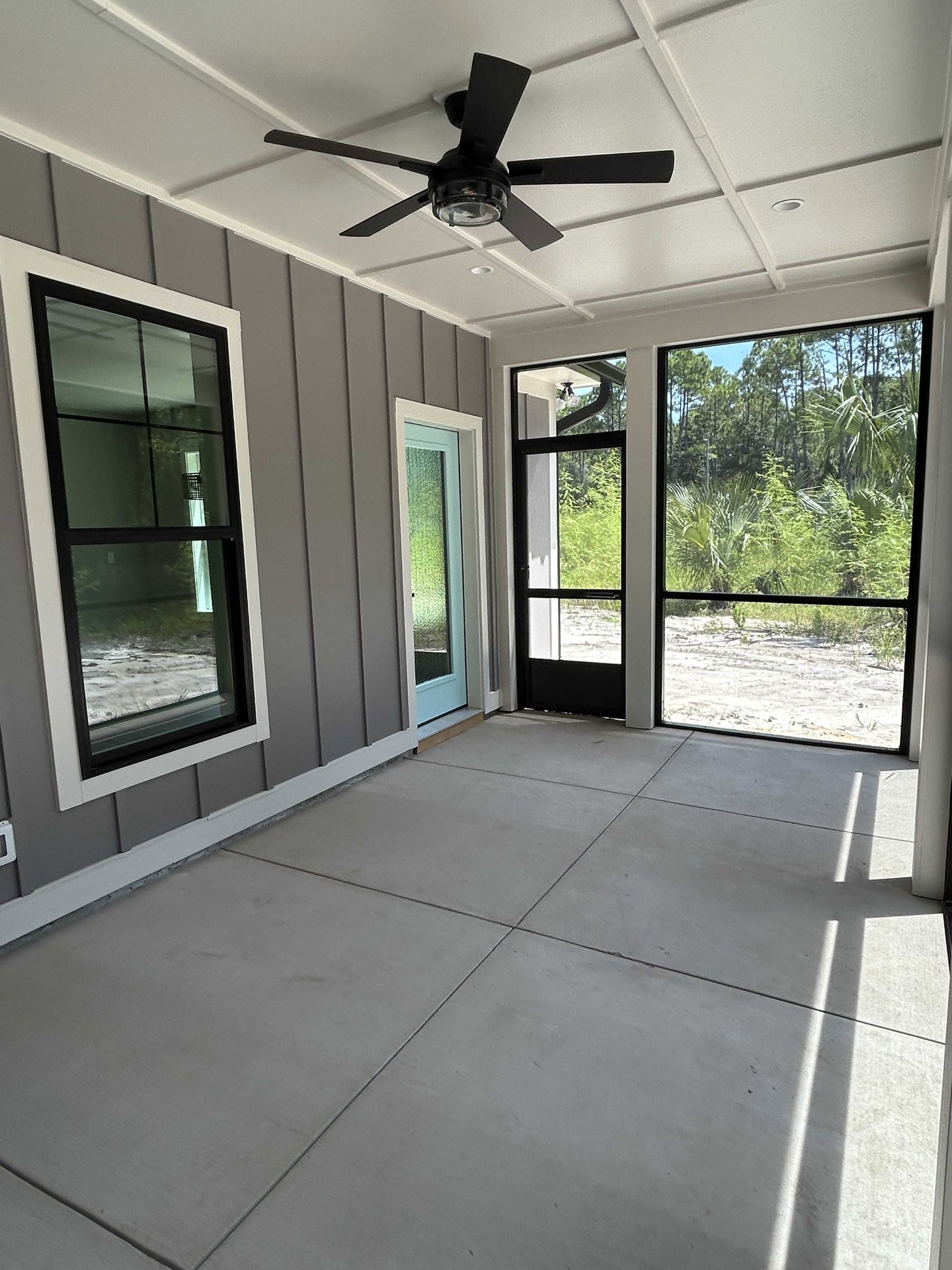 Ceiling fan with glass light fixture above concrete floor, black-framed window and door opening to view of trees and sandy landscape