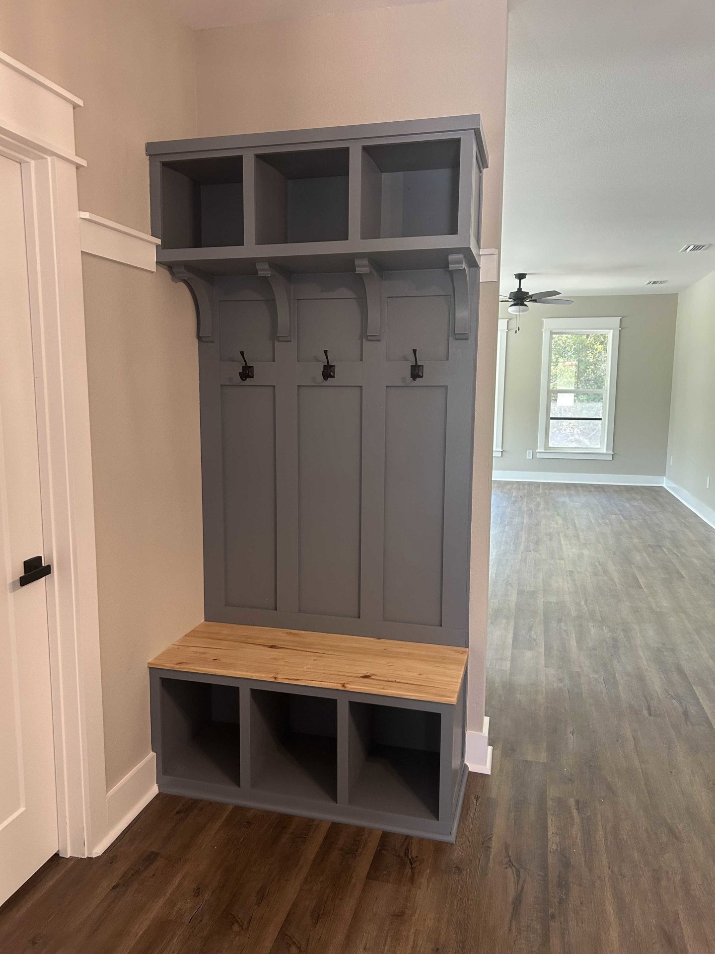 Hallway with wood flooring, white door with black handle, grey wall-mounted coat rack with hooks, shelf featuring wood top, window with white frame.