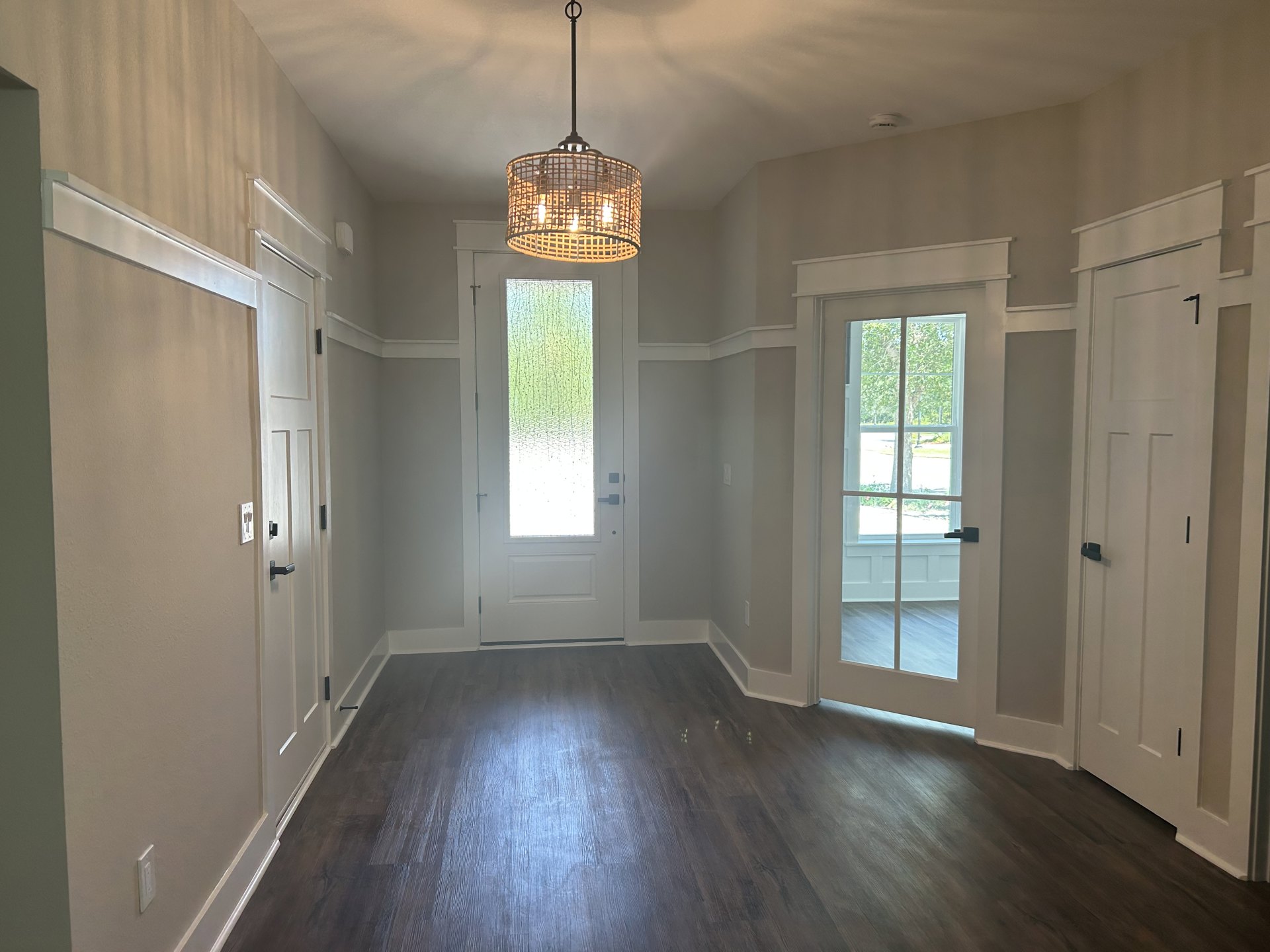 Hallway with wood flooring, basket-style chandelier on ceiling, glass-paneled door open to reveal tree outside, window close-up, white walls with molding
