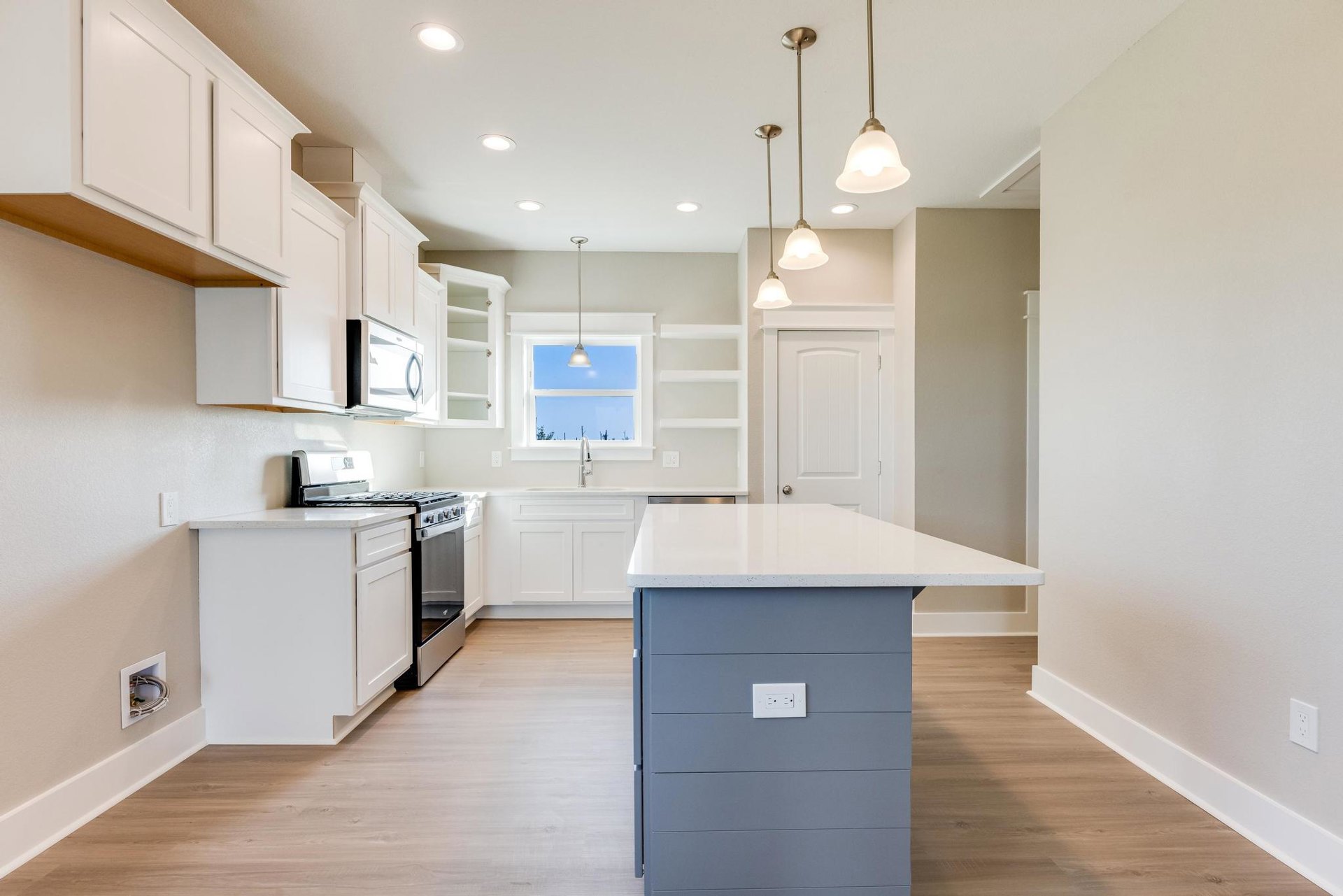 White and grey kitchen island with built-in stove and oven, white cabinetry, black button outlet, window letting in natural light, light wood flooring, and visible plumbing pipe