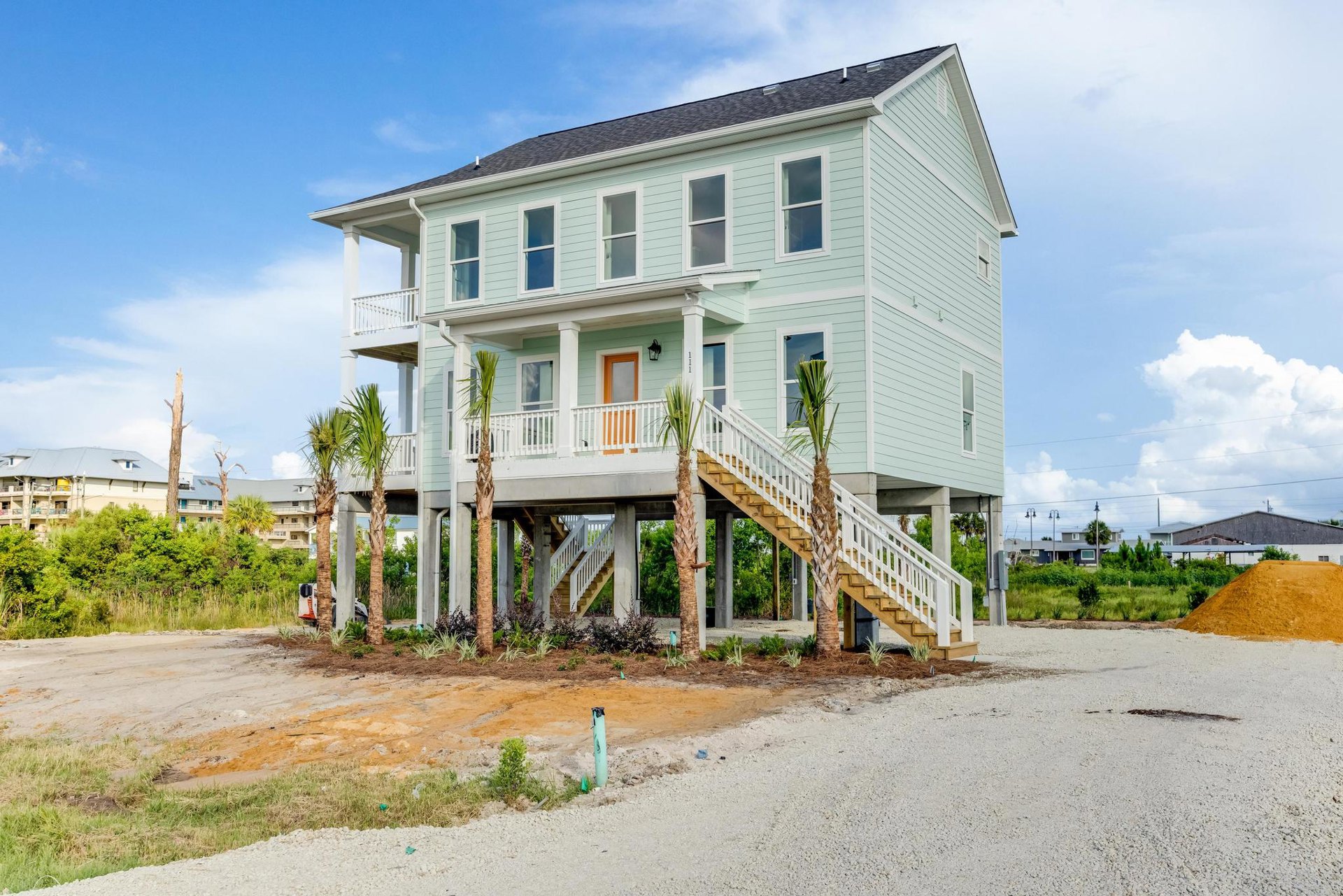 Modern two-story house surrounded by mature palm trees, white-framed windows, outdoor staircase, sandy yard with scattered dirt piles, and cloudy sky overhead