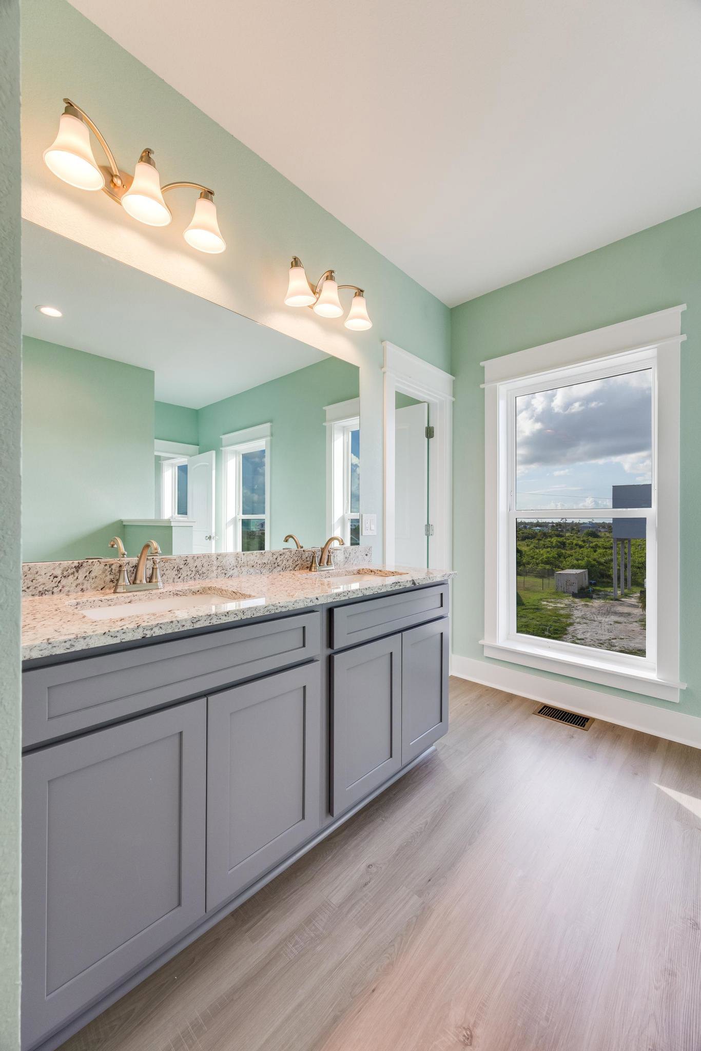 Bathroom with double sinks, expansive mirror, light fixture with three bulbs above, window overlooking a field and building, grey cabinetry, wood flooring, and wall-mounted lamp.