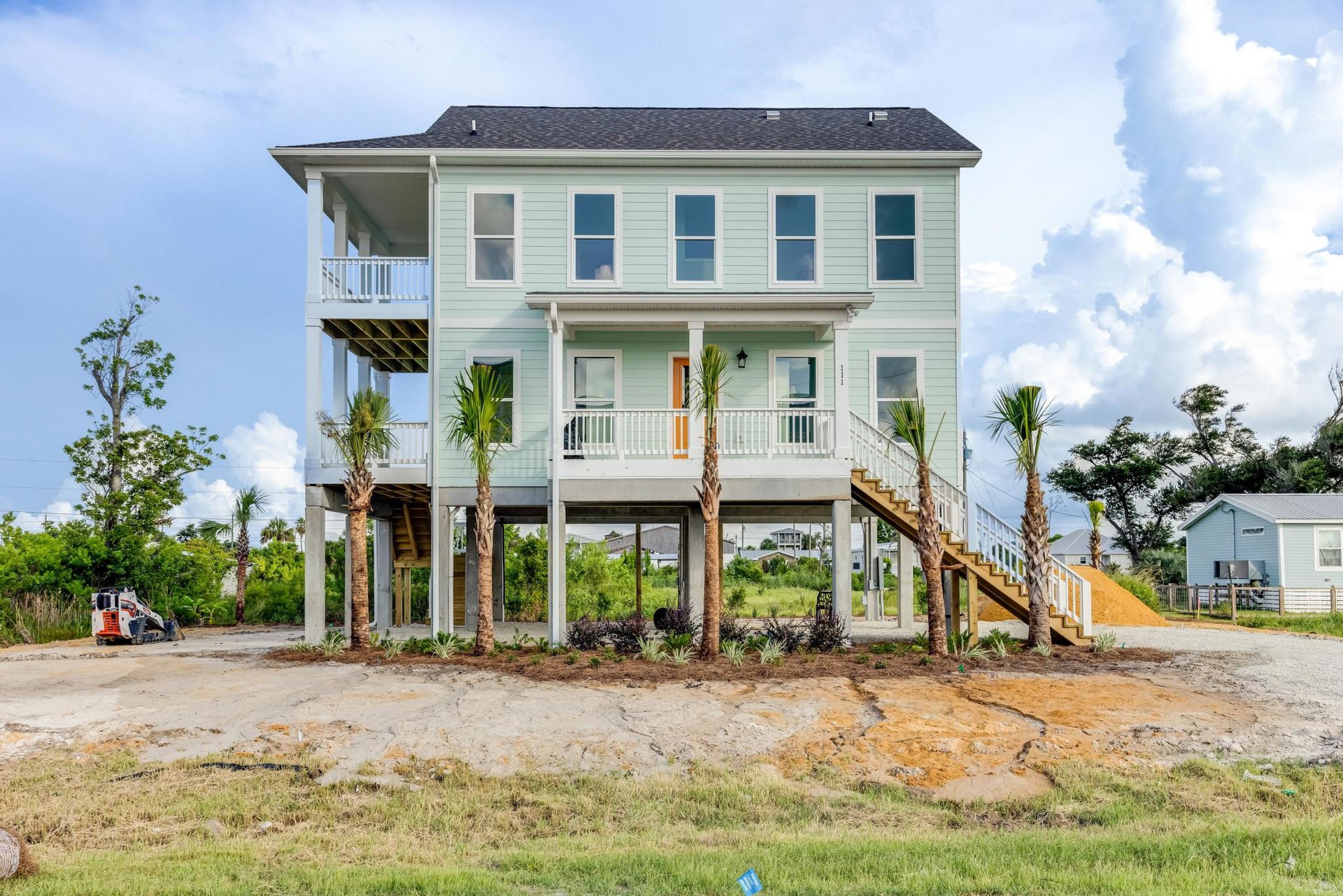 Modern home with white-framed windows, blue glass panels, and a covered porch, surrounded by palm trees, grass, and a dirt road; white and orange construction equipment visible