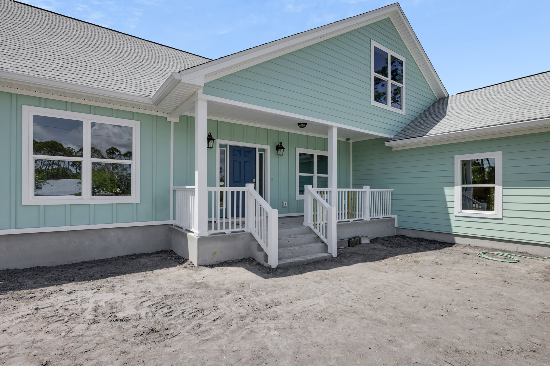 Blue front door and white porch with railing, white-framed windows reflecting trees, exterior siding, dirt and stairs leading to entrance