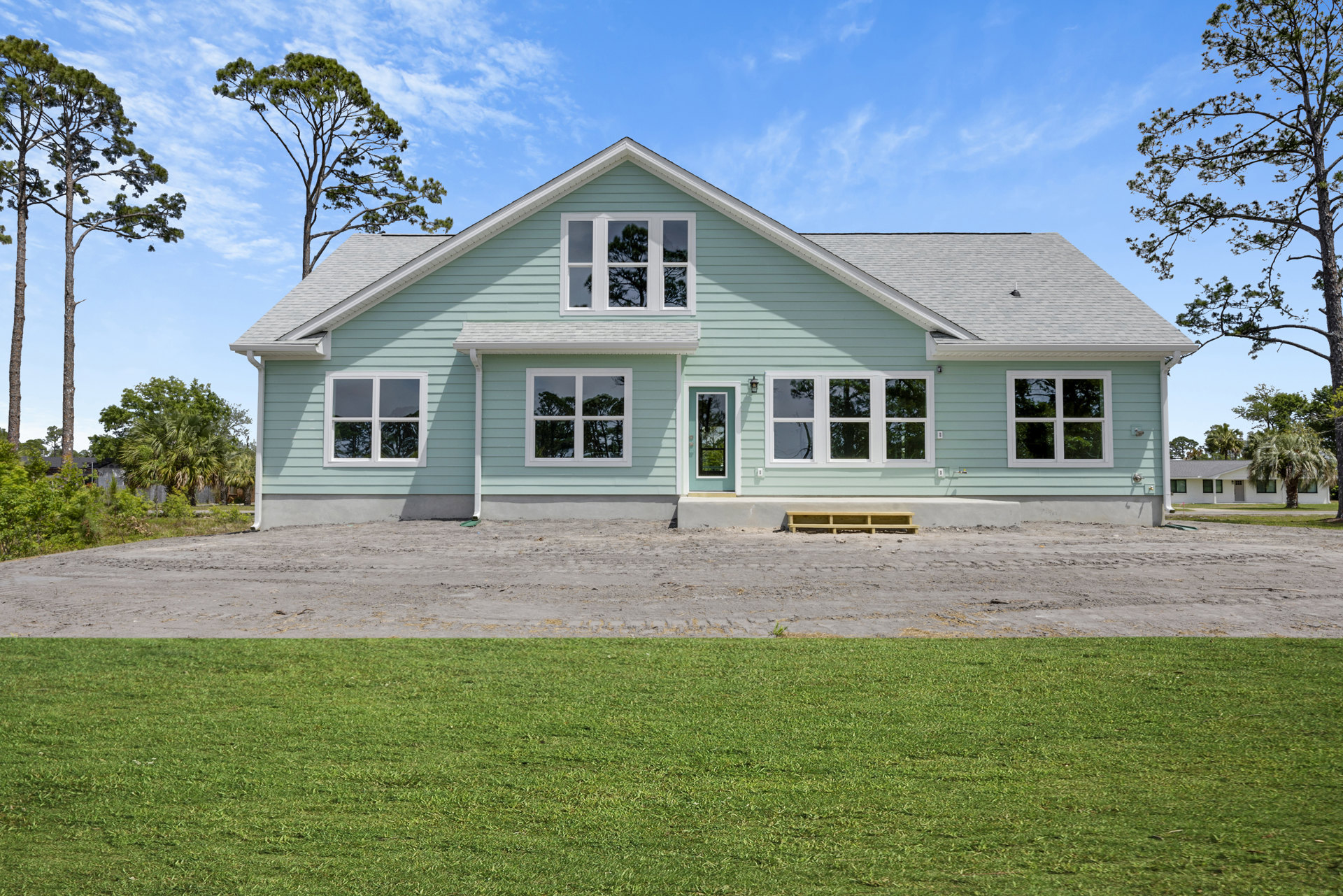 Blue siding house with white trim, large windows reflecting nearby trees, expansive green lawn bordered by a stone wall, under a partly cloudy sky