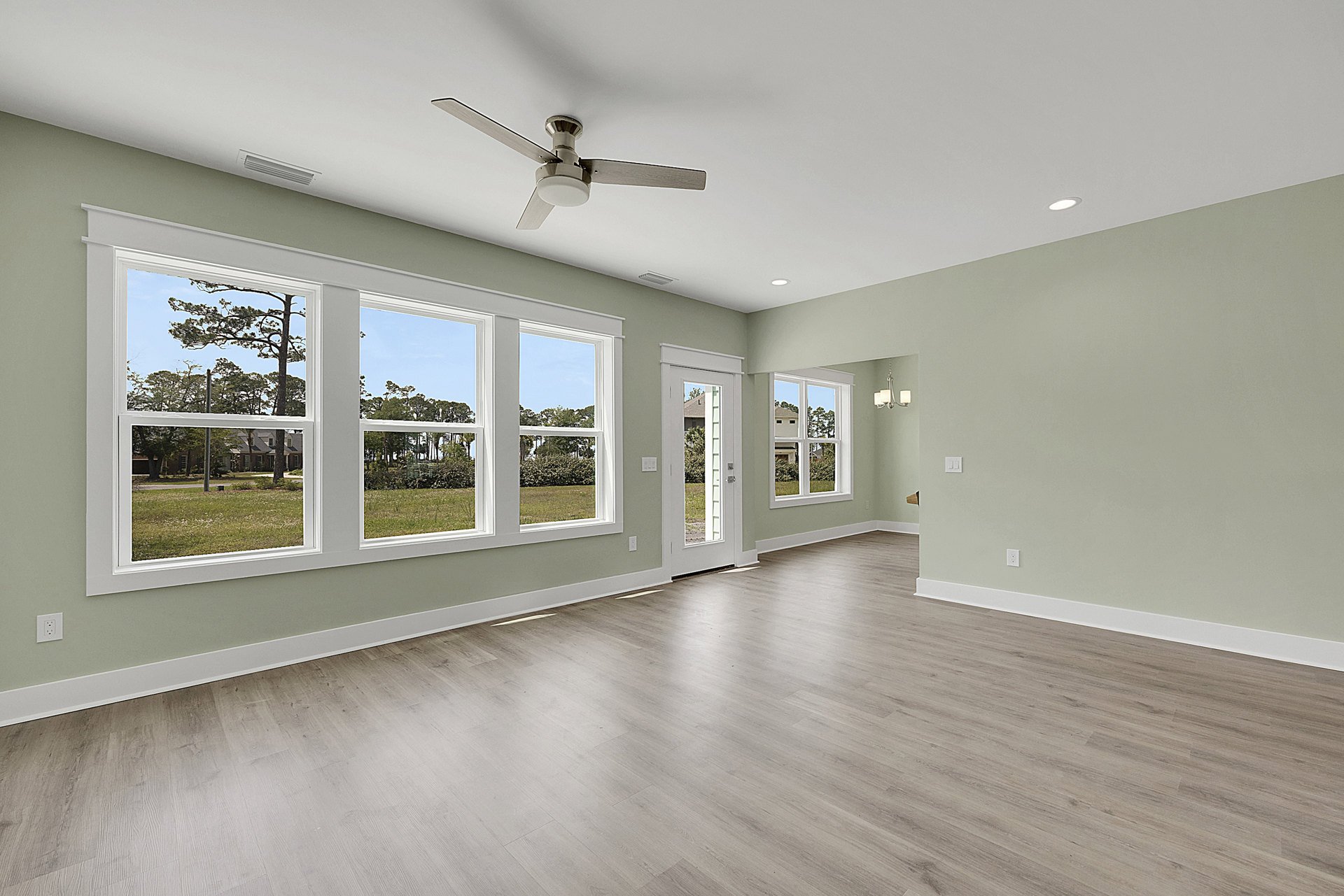 Living room with laminate wood flooring, white plaster walls, ceiling fan with light, large windows overlooking green lawn and trees.