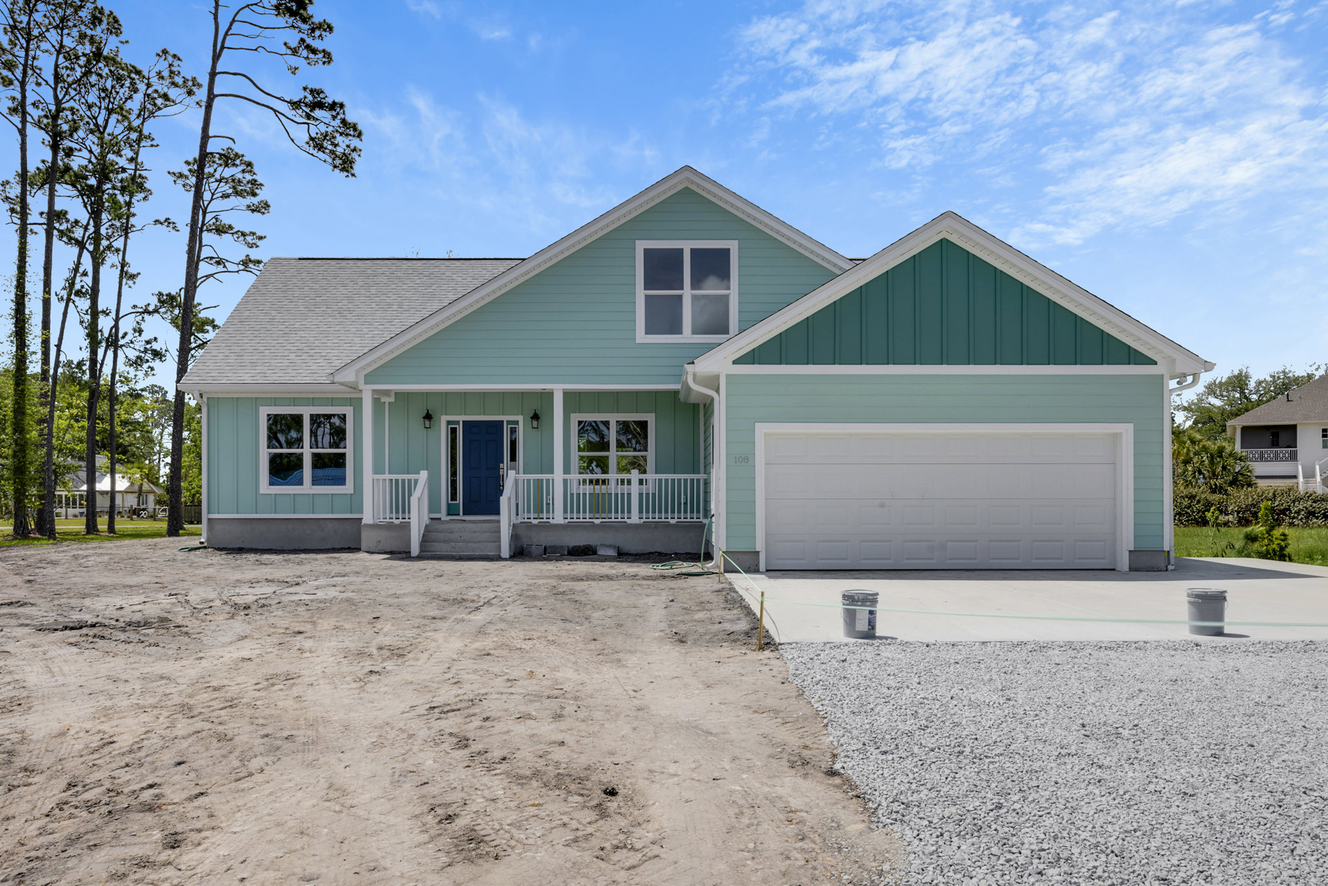 Two-story house with white porch railing, multiple windows reflecting clouds and trees, grey trash can and bucket near driveway, cloudy sky above, trees in background.