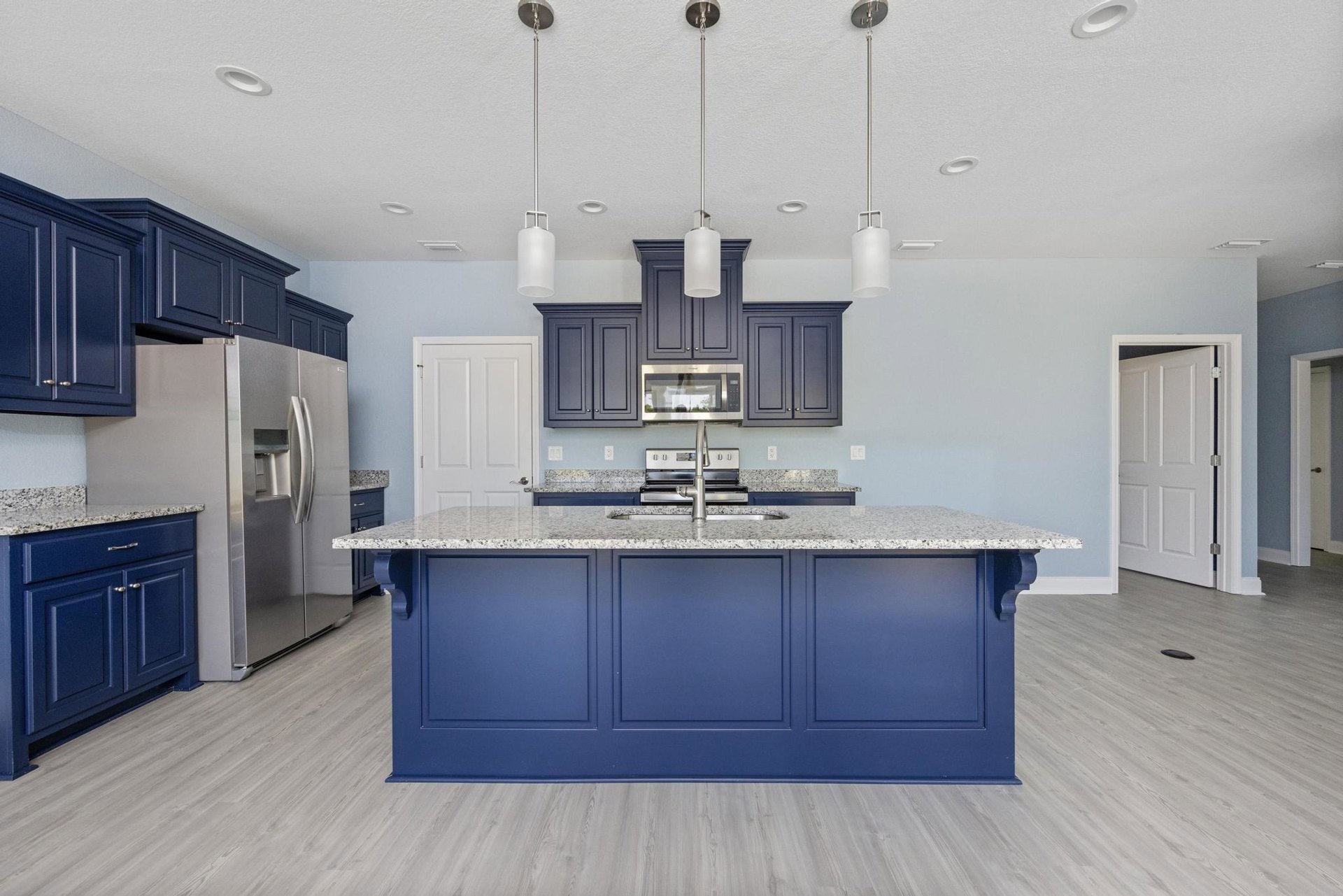 Spacious kitchen featuring a large central island with stone countertop, white cabinetry, stainless steel refrigerator, undermount sink, white door with black handle, black-trimmed