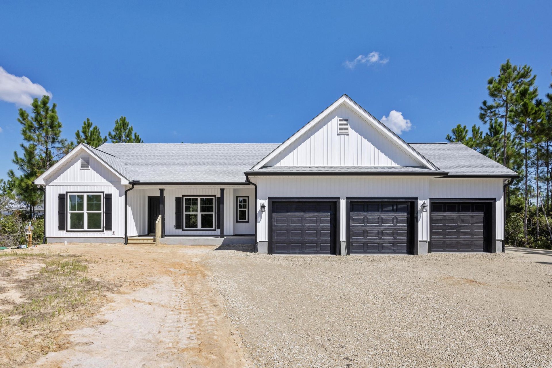 Two-story home with white siding, black shutters, and white-trimmed windows; attached garage with paneled door; paved driveway bordered by trees; vented roof under partly cloudy