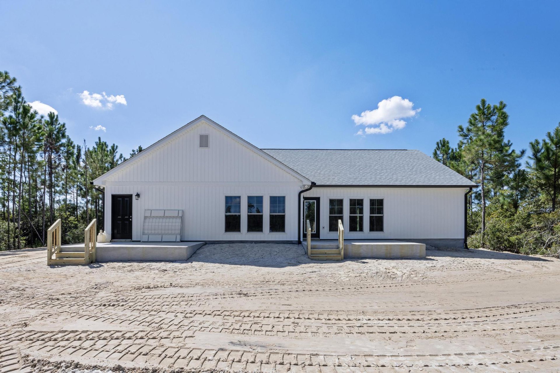 White two-story house with wooden railing, sandy driveway showing tire tracks, white cloud in blue sky, wooden stairs with handrail, black grid shelf visible near entrance.