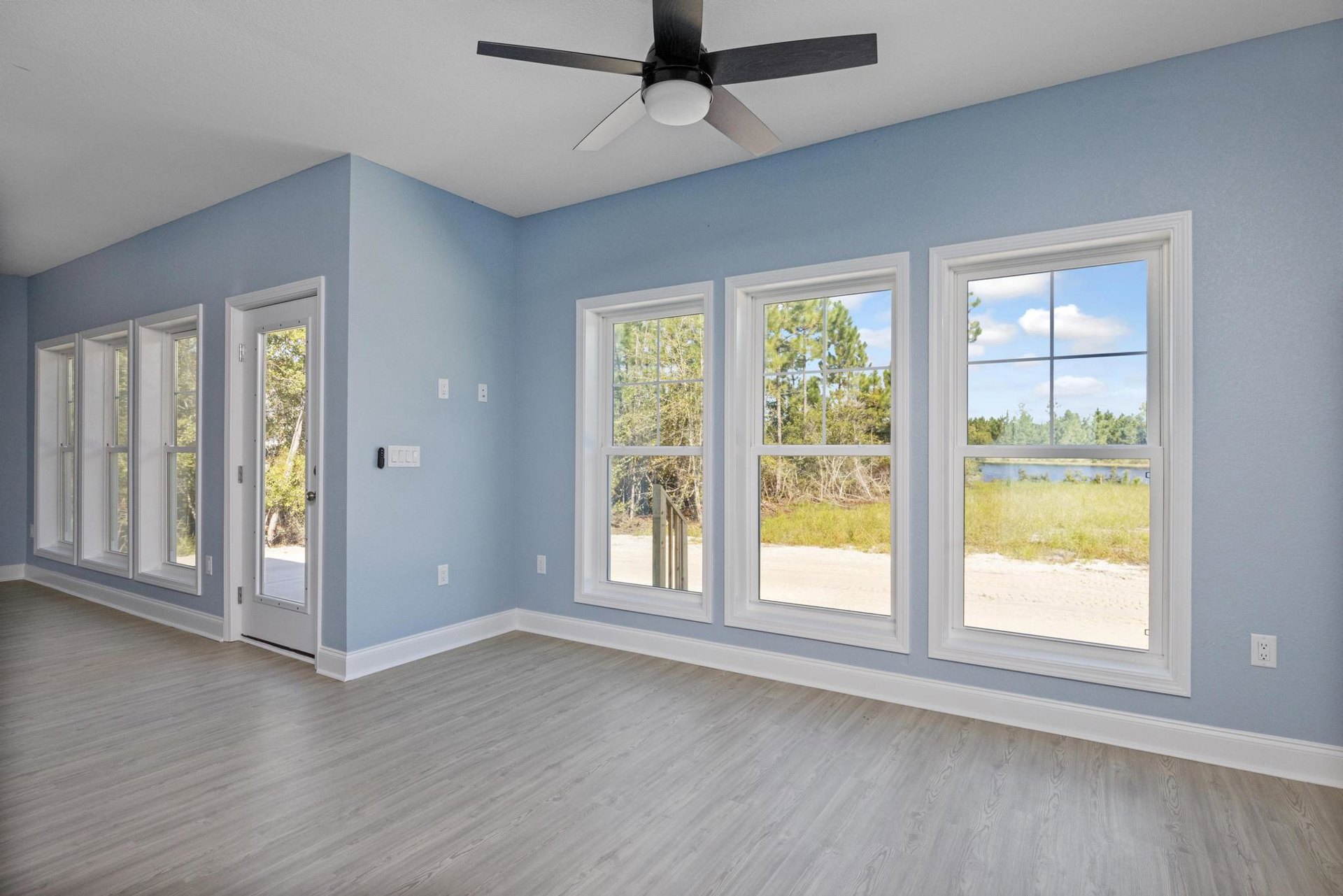 Ceiling fan with light fixture above wood flooring, multiple windows showing views of trees, river, and open field