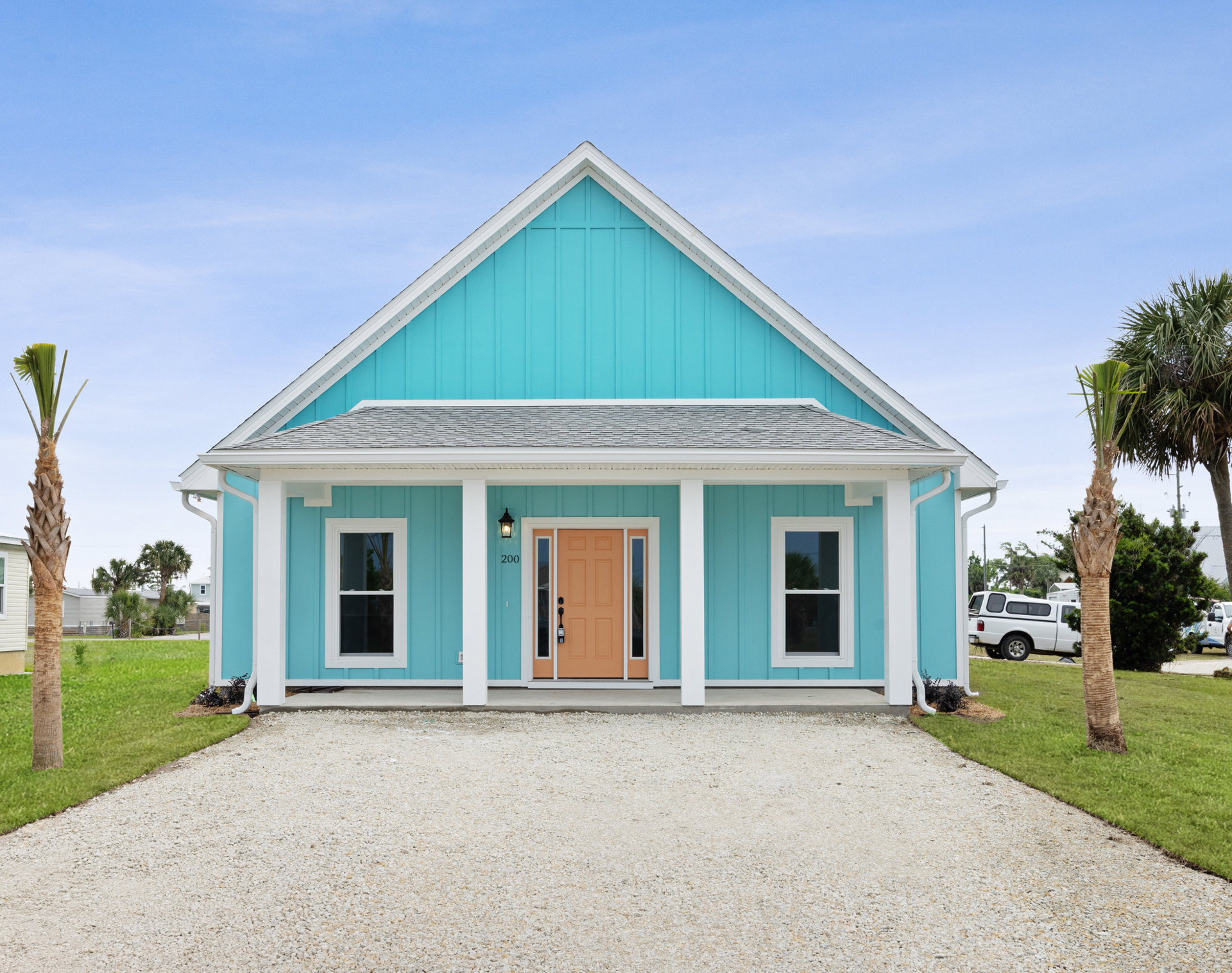 Blue siding house with yellow front door, gravel driveway, white truck parked nearby, palm tree visible through window, cloudy sky overhead.