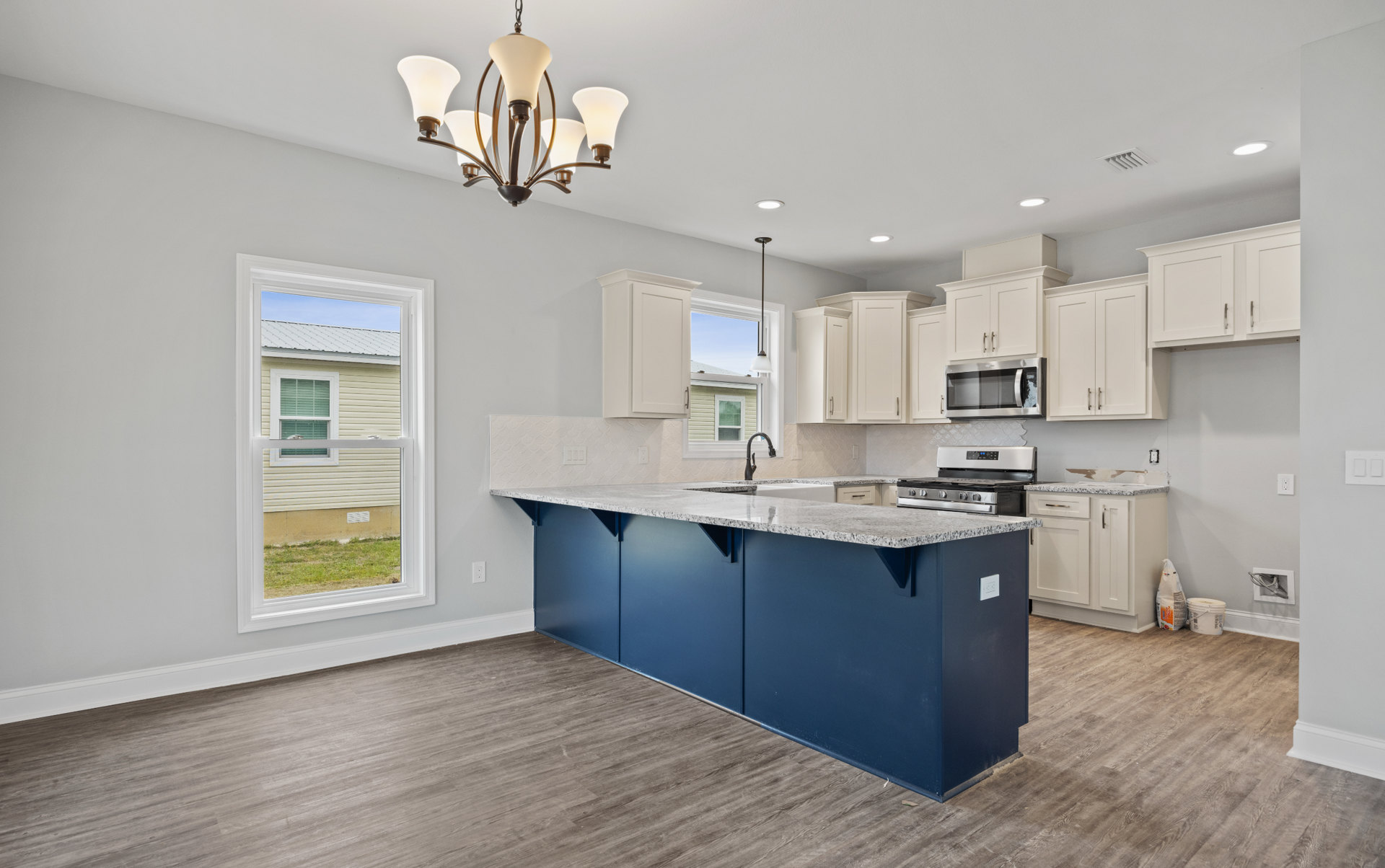 Blue kitchen island with white cabinets, white quartz countertops, black faucet, stainless steel microwave, window with white trim, and modern chandelier