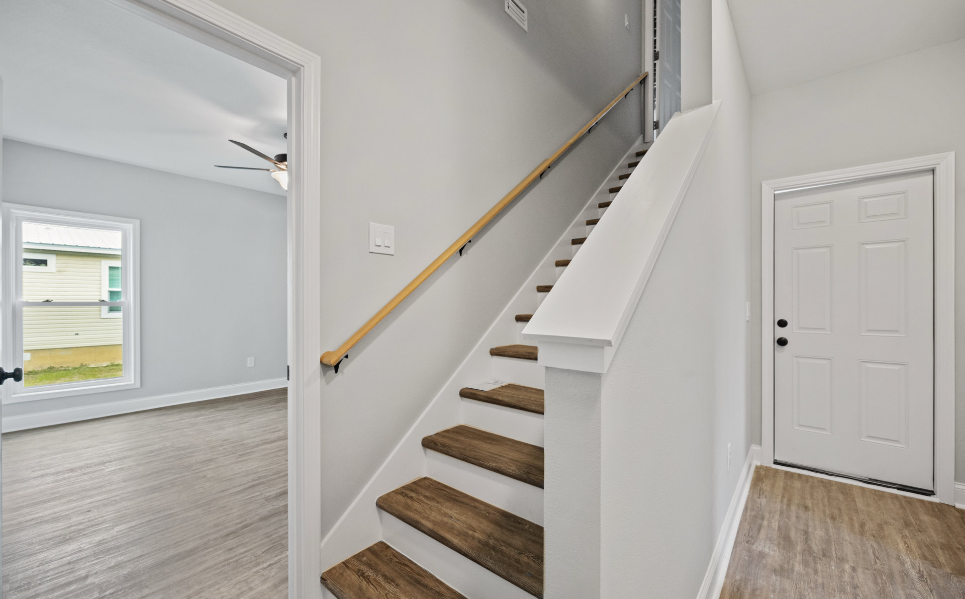 Wooden staircase with white risers and black handrail, white door with black hardware, large window overlooking green lawn, light hardwood flooring, smooth plaster walls