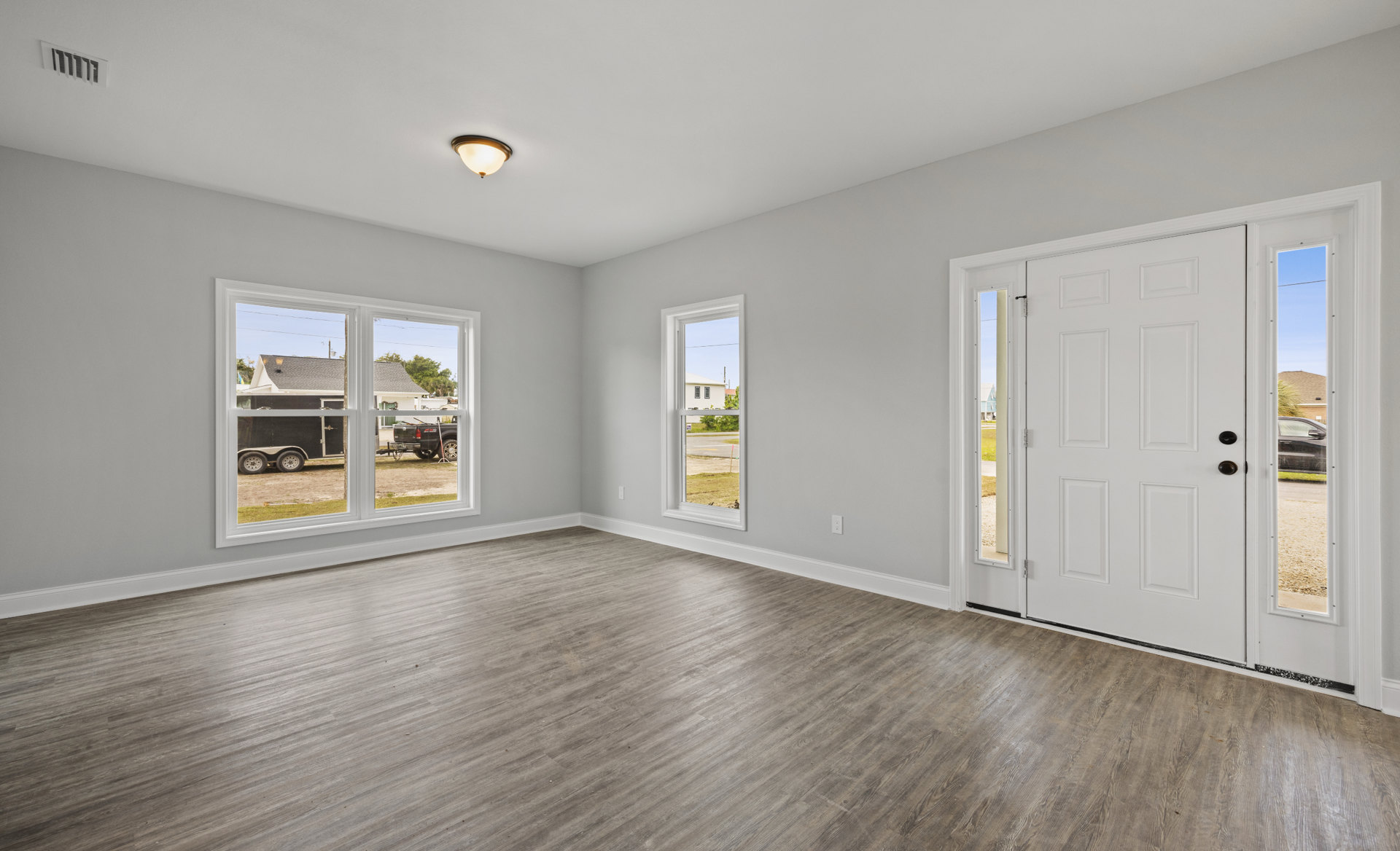 Room with light wood flooring, white plaster walls, large windows, and a paneled door; ceiling features recessed lighting.