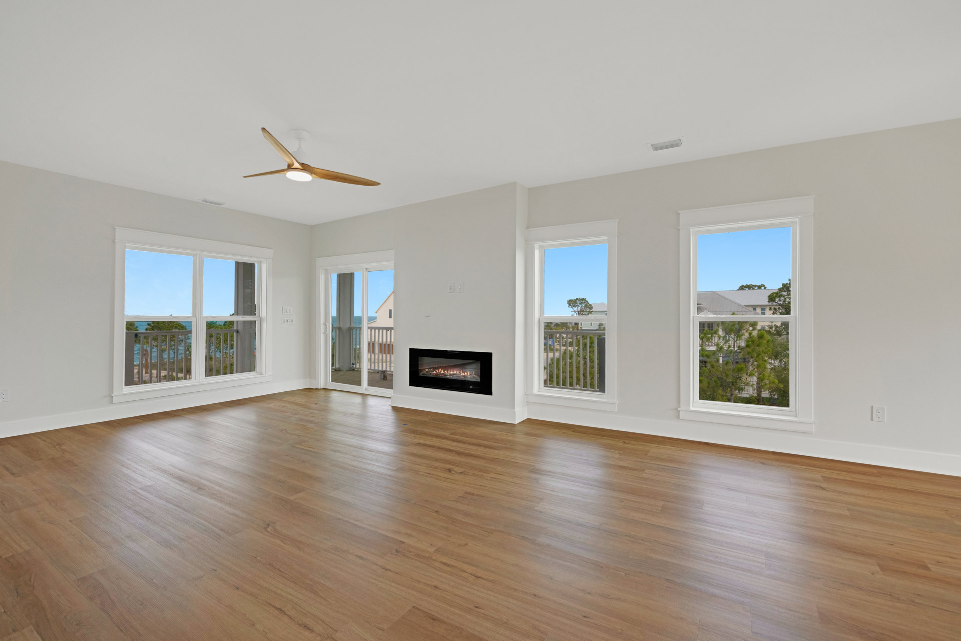 Living room with hardwood floors, stone fireplace with burning fire, ceiling fan with light, large windows showing trees and ocean view