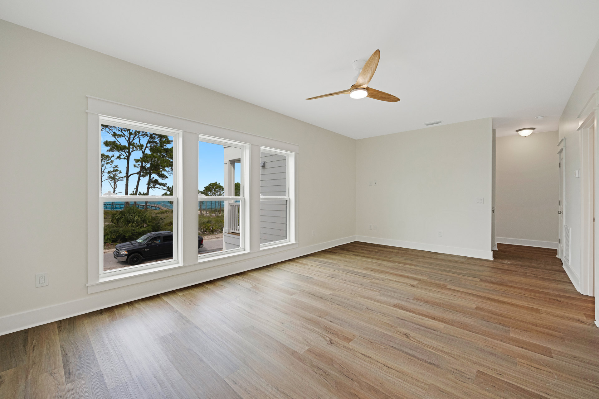 Living room with wood flooring, ceiling fan with light, large windows showing trees outside, white plaster walls