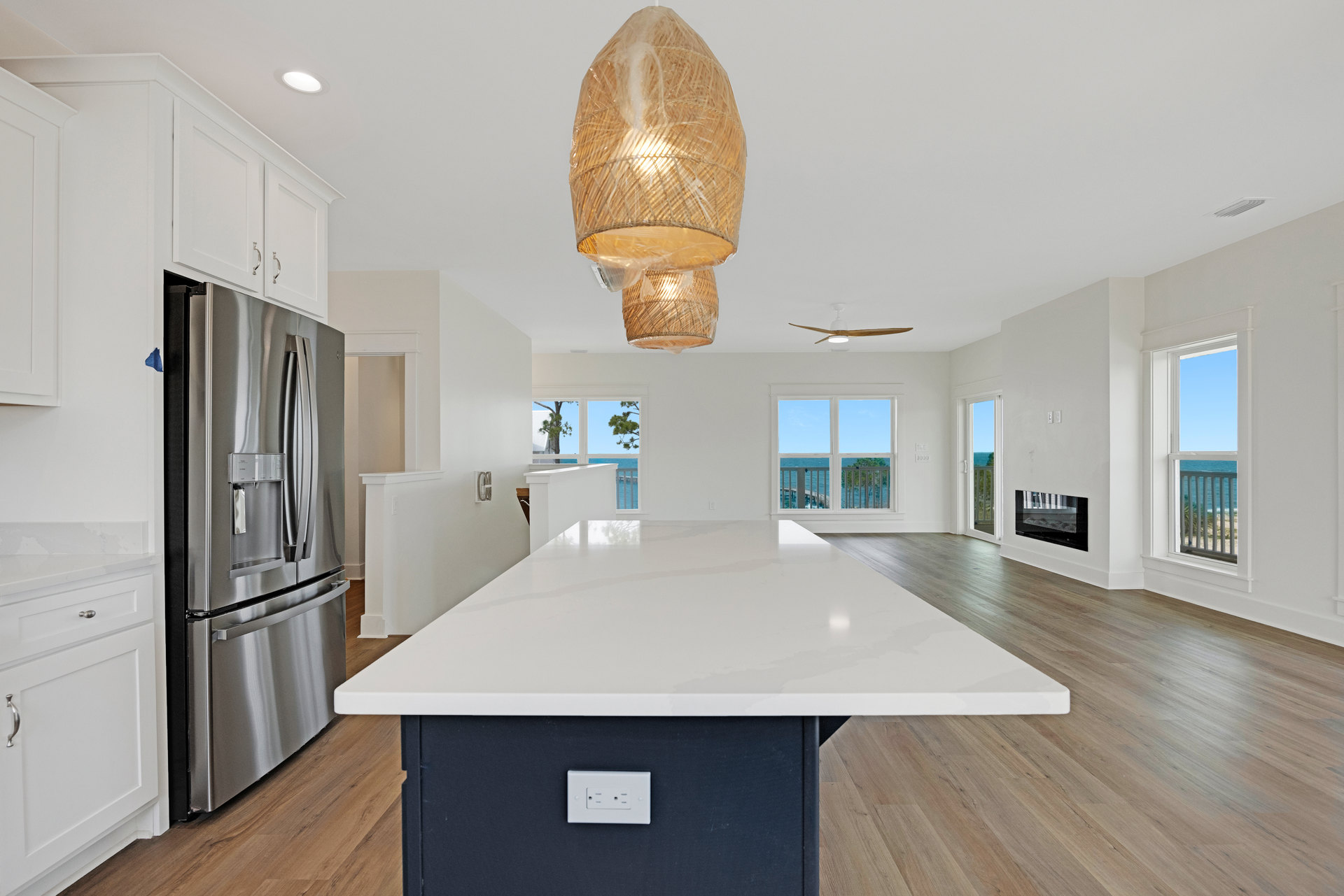 Open-concept kitchen featuring a central island with quartz countertop, oversized pendant light wrapped in protective plastic, stainless steel refrigerator, white cabinetry, and