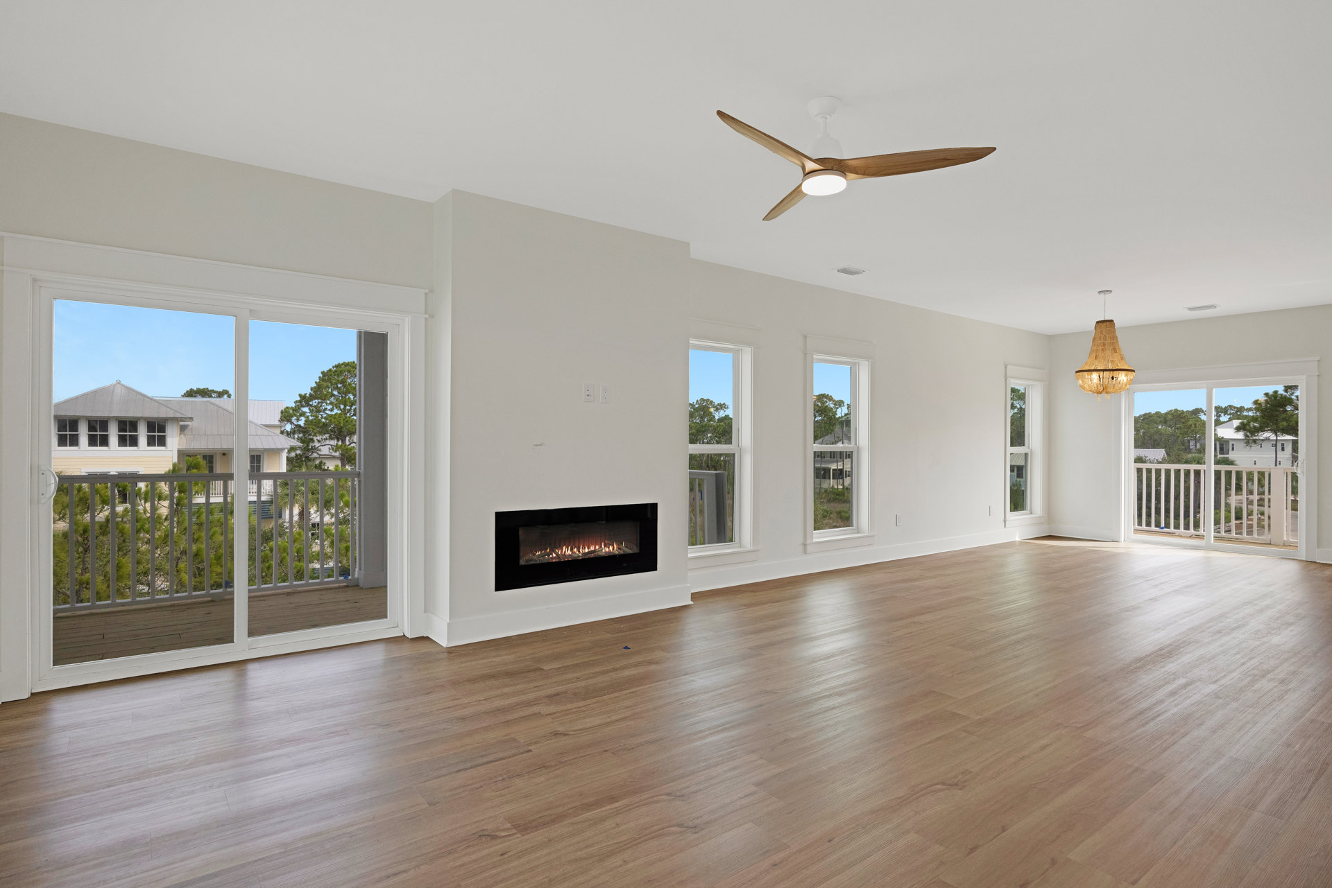 Spacious living area featuring wood laminate flooring, stone fireplace with active fire, ceiling fan with integrated light, and partial view of wood railing on adjacent deck