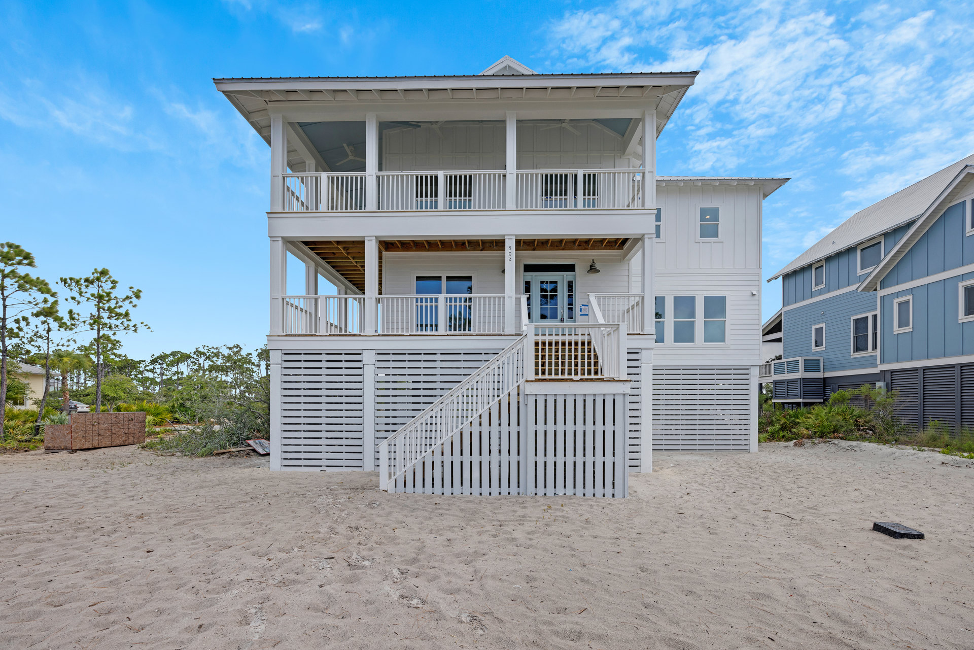 White beachfront house with wide deck featuring white railings, sandy yard bordered by a white fence, large windows, and blue sky overhead