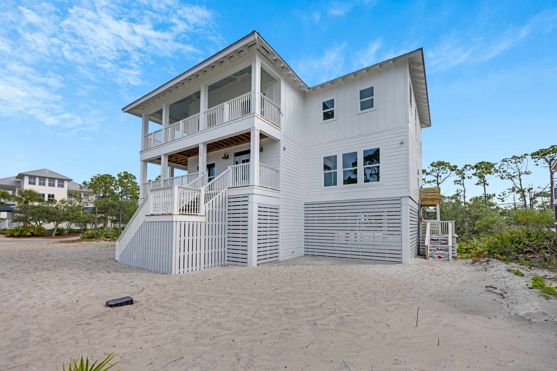 White beachfront house with sand and wood fence, large windows, and porch; staircase visible with white plastic bags and cement bags nearby; cloudy sky and trees in background.